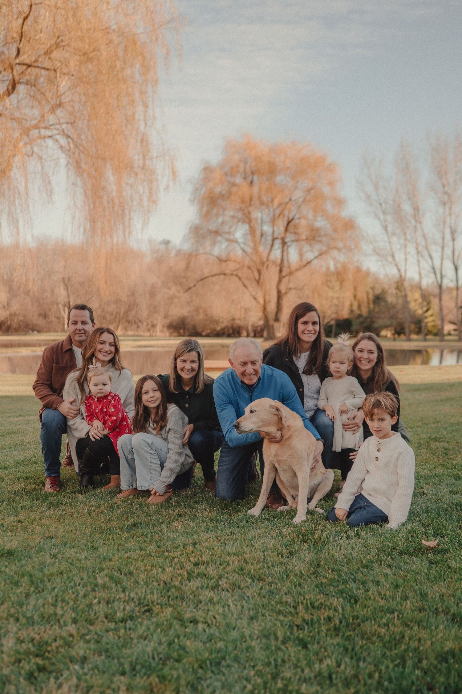 Extended family gathers for outdoor portrait with their yellow lab on grassy field during golden hour.