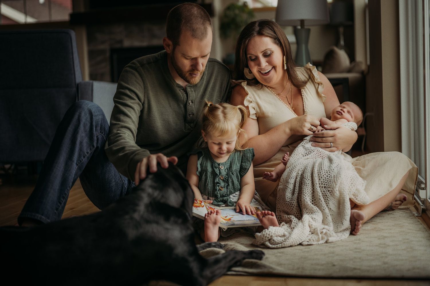 Family gathering with black dog, toddler and infant sharing a quiet moment by window.