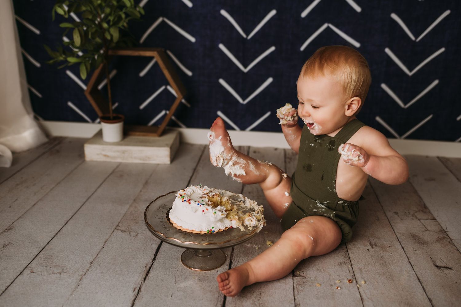 Small child in olive romper sits on wooden floor with cake during messy cake smash photo session.