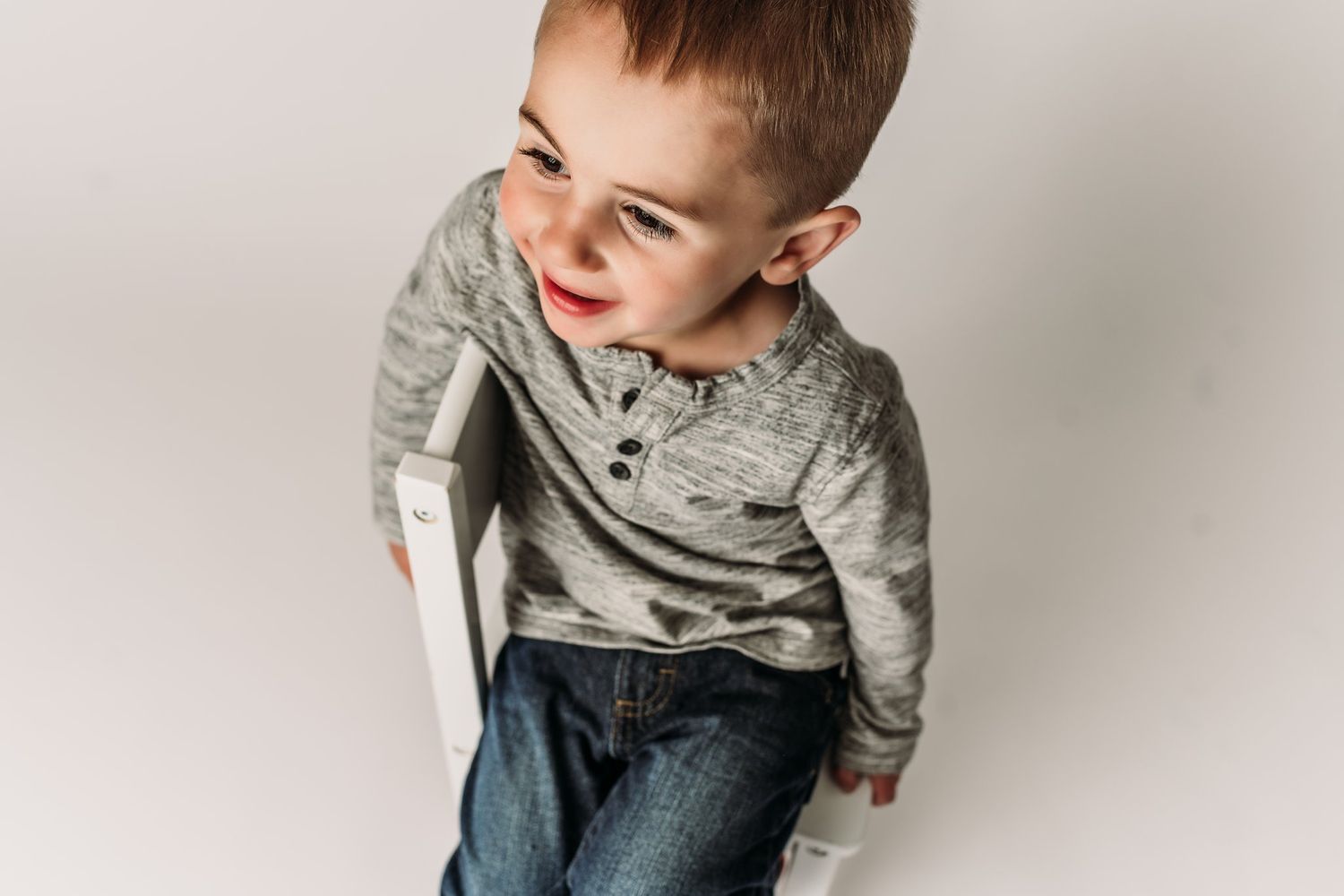 Child wearing gray henley and jeans poses candidly against white background.