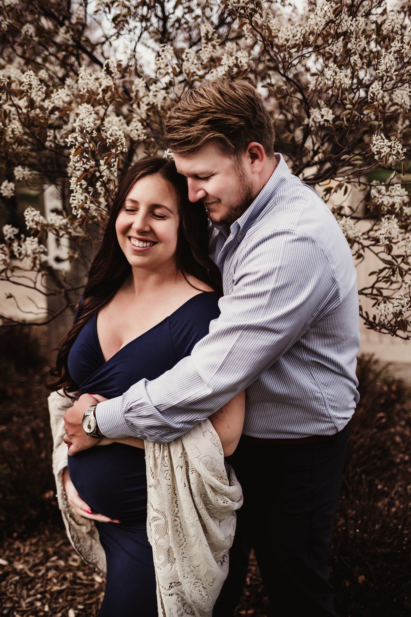 Tender moment captured between expectant couple against blooming spring trees.