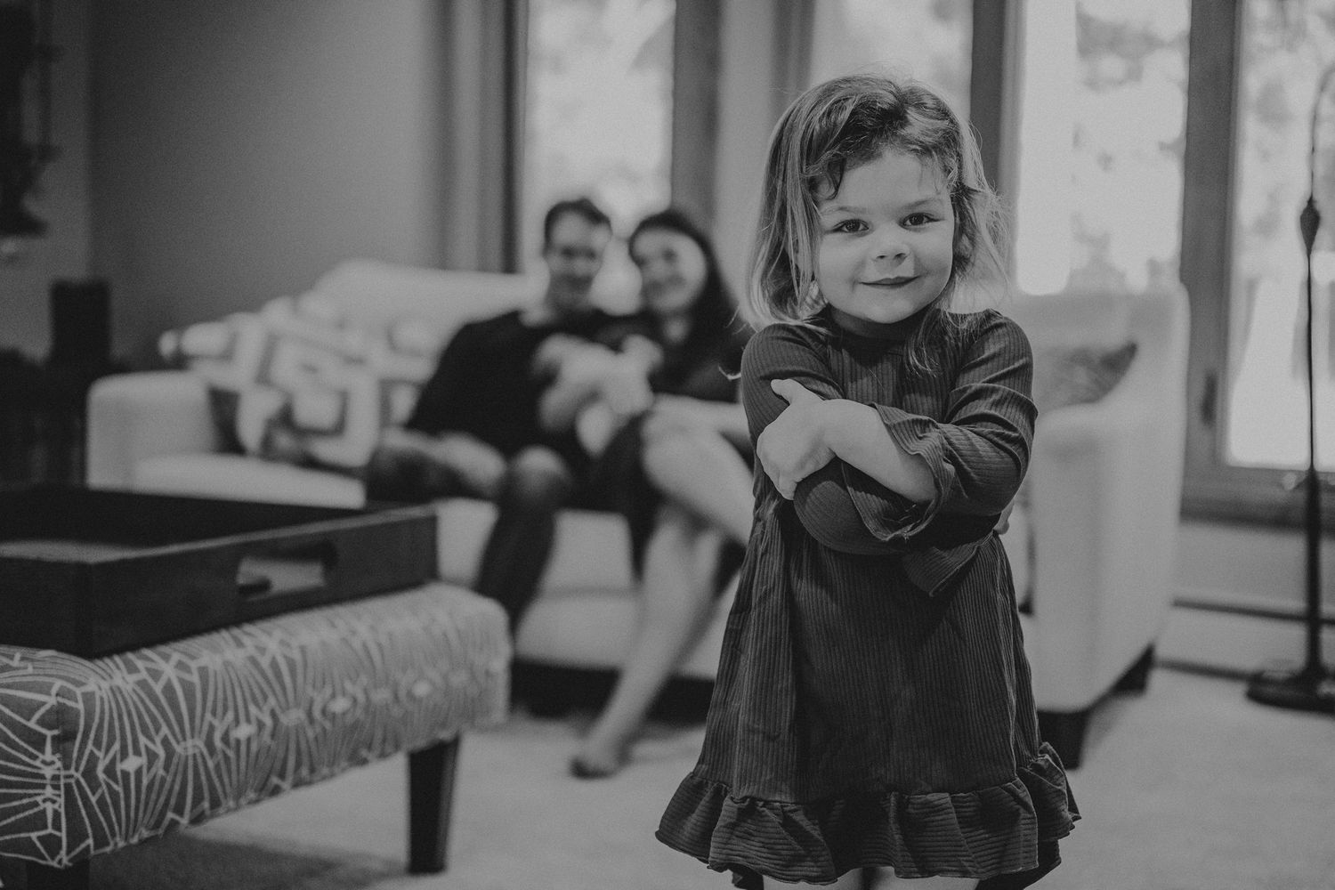 Black and white photo of a toddler in dress standing in foreground with family on couch in background.
