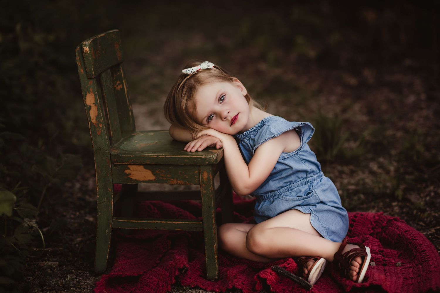 A child rests their head on a vintage green chair atop a red blanket in a forest setting.