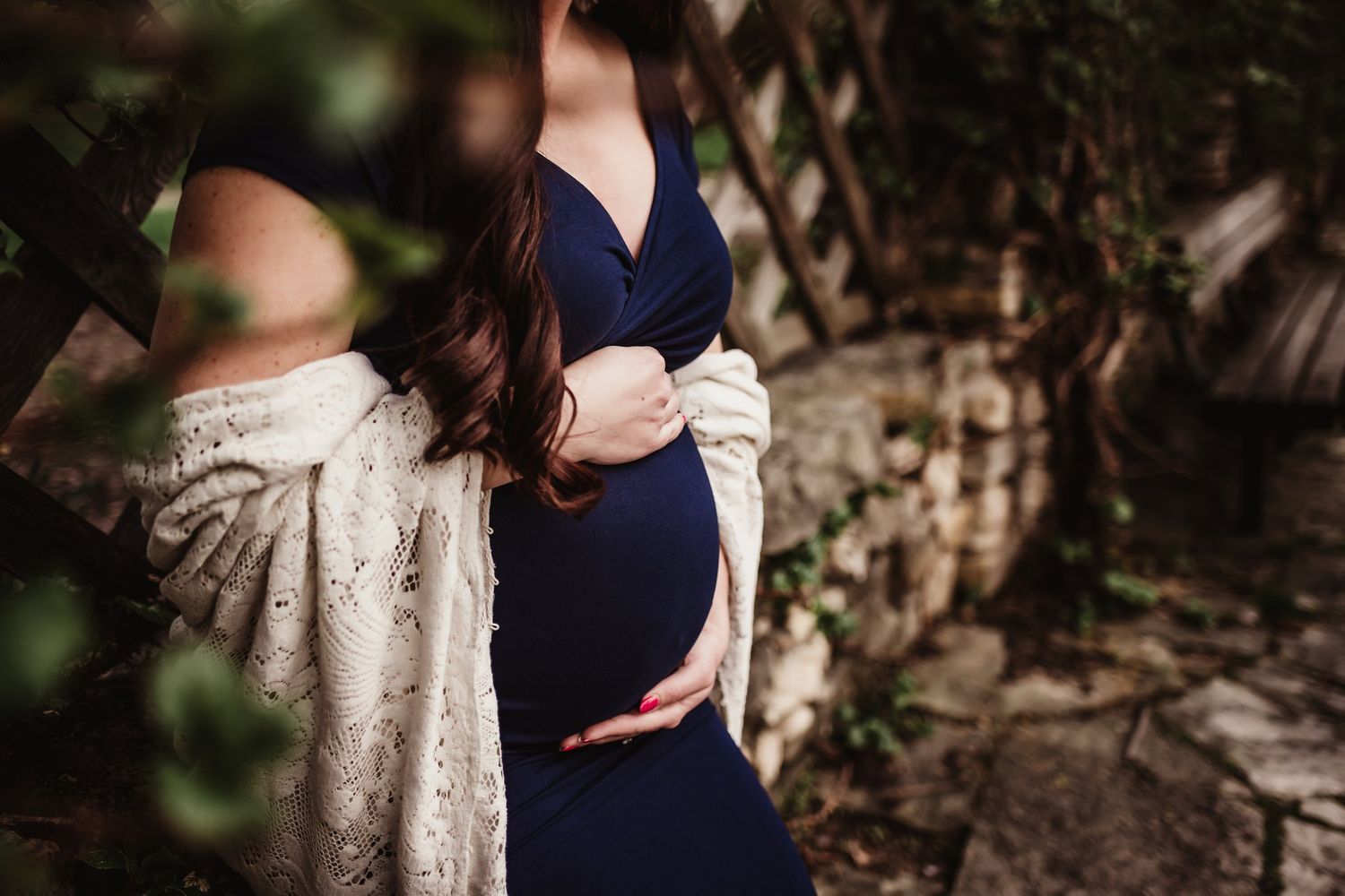 Artistic maternity photo showing woman in navy dress against stone wall with lace cardigan.