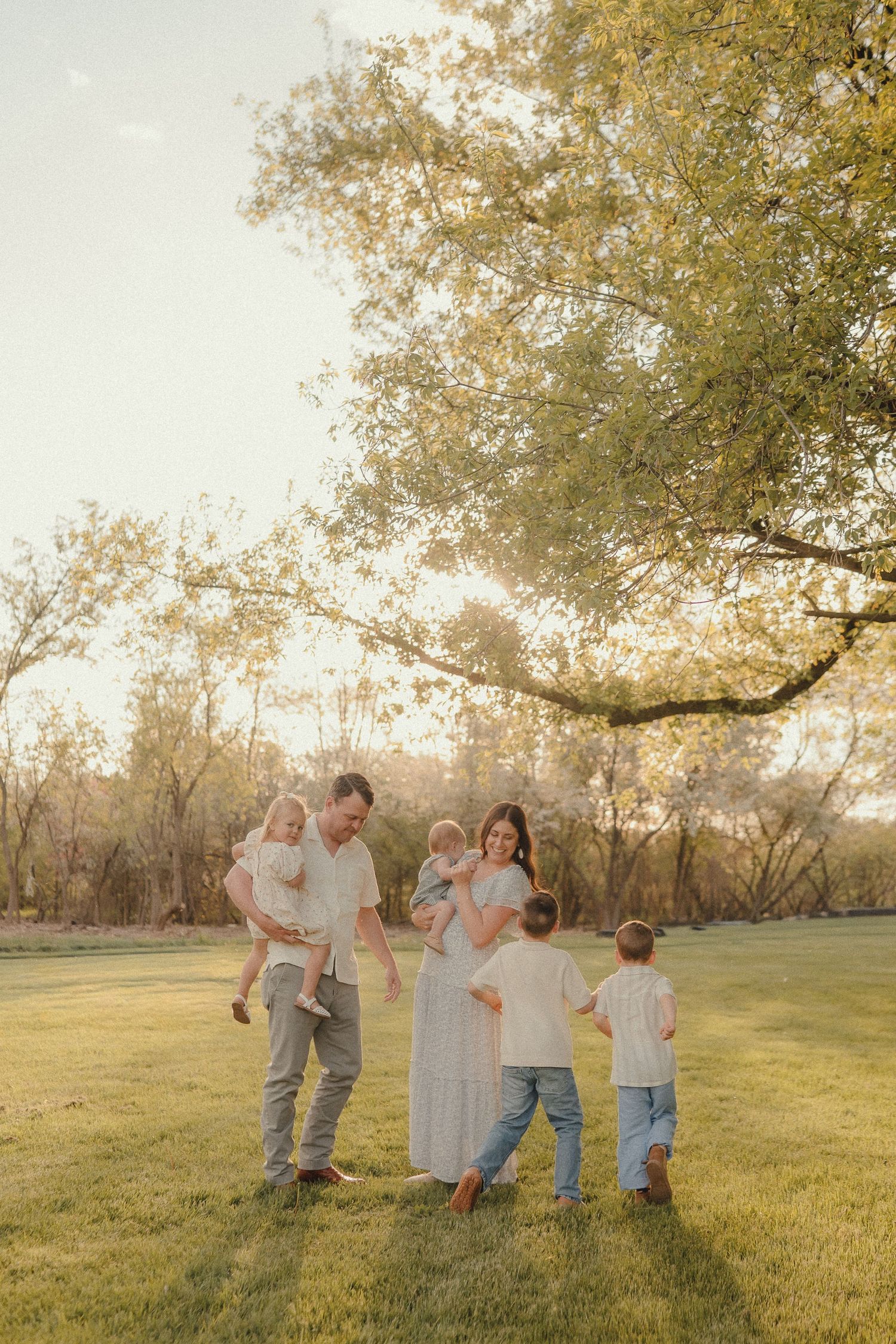 Family of six enjoying golden hour outdoors, parents holding toddlers while older children play on lush green grass under large tree.