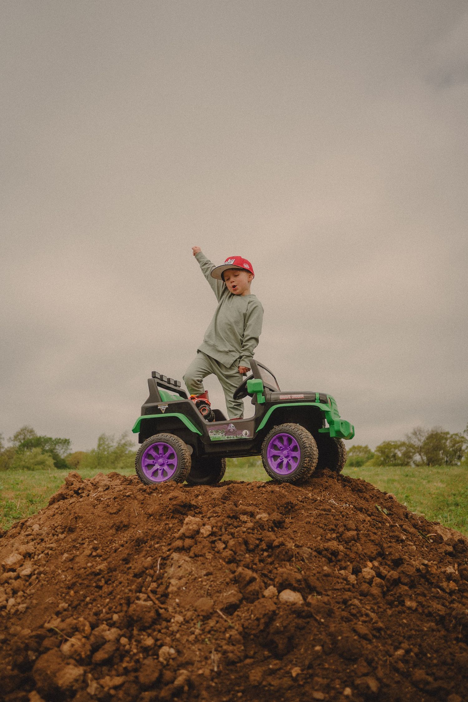 Adventurous boy in red helmet stands triumphantly atop a dirt mound on a green toy electric jeep.