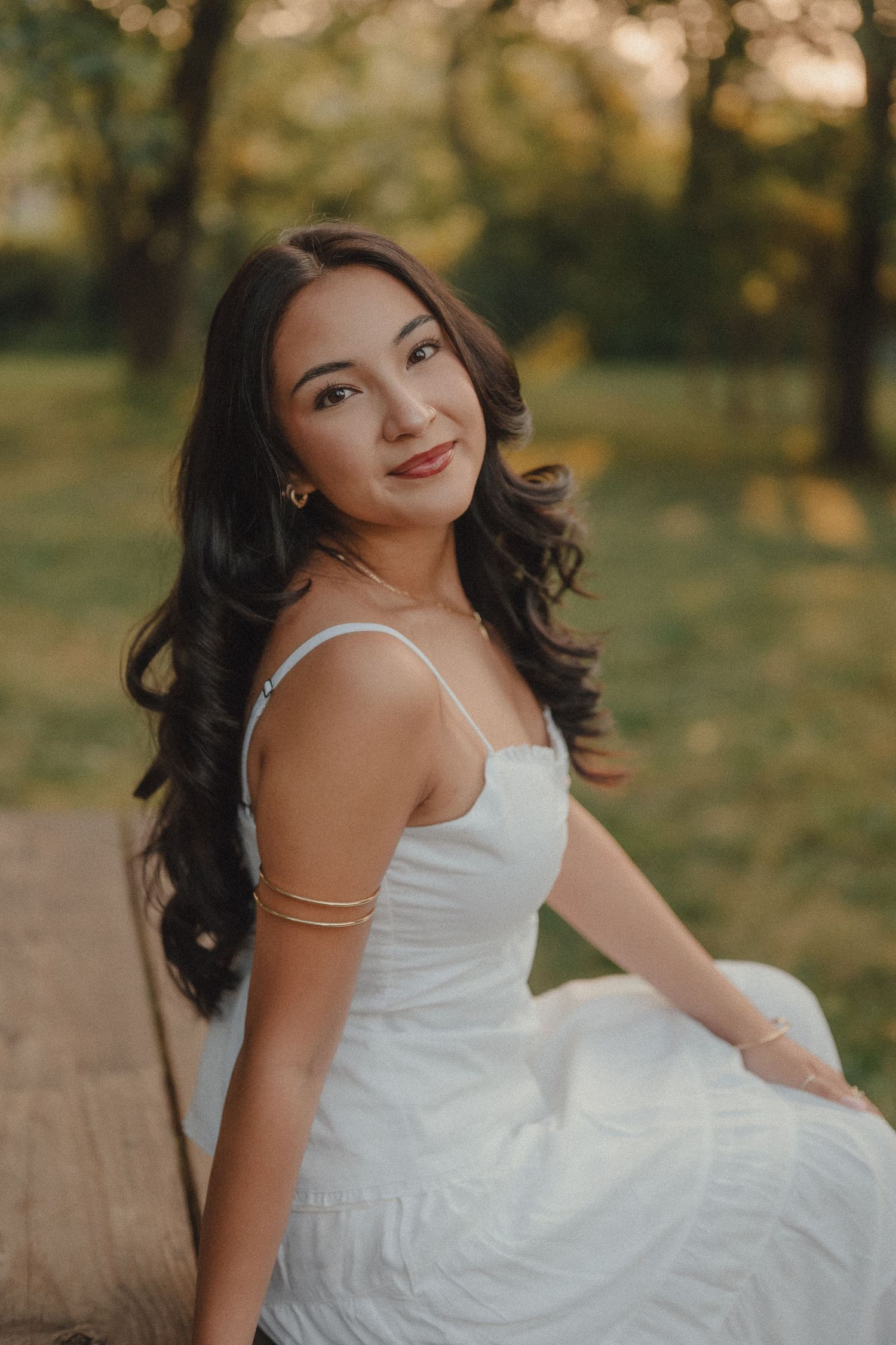 Young woman in white dress sitting on bench in park at golden hour, wearing gold jewelry.