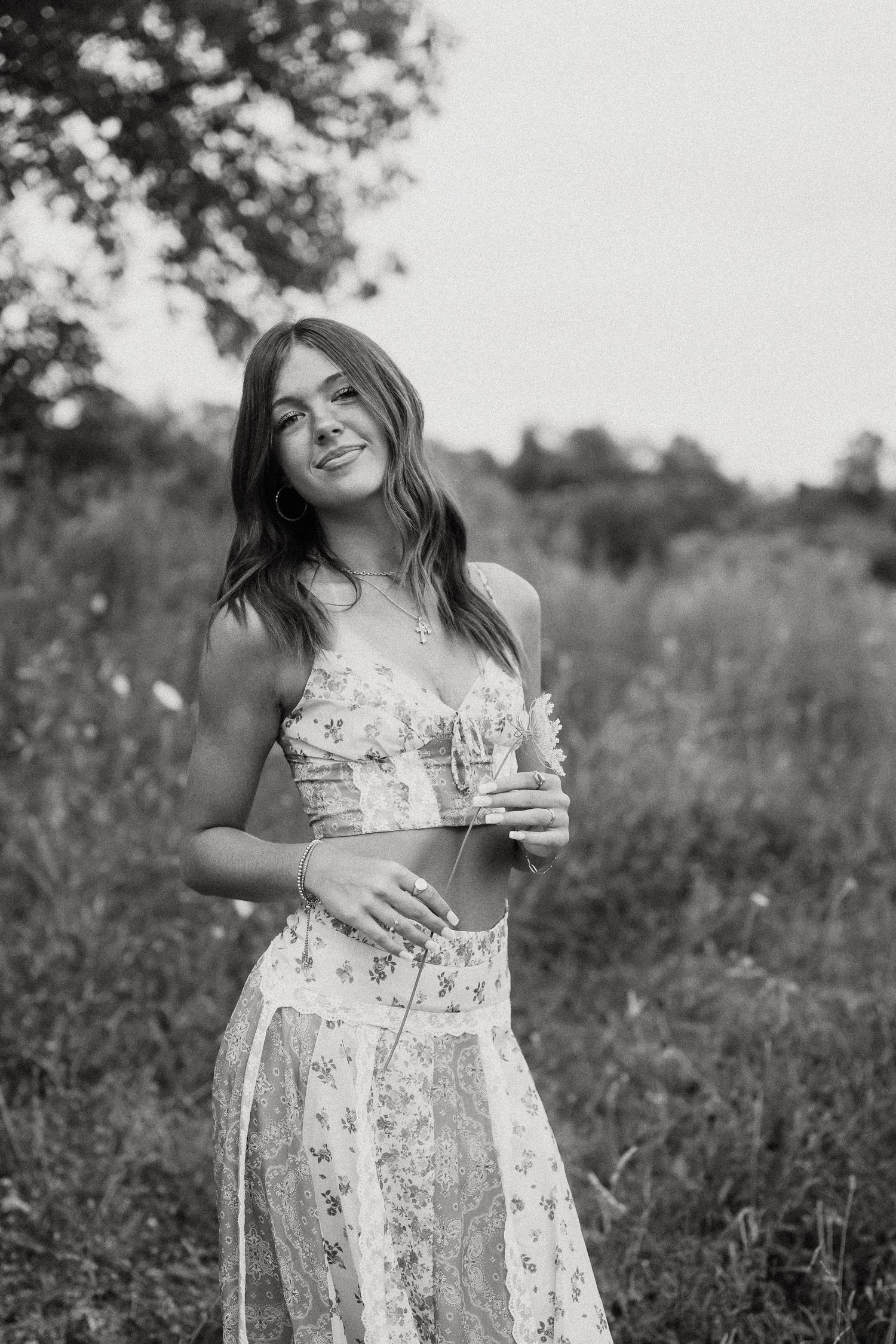 Black and white portrait of young woman in floral crop top holding wildflowers in grassy field.