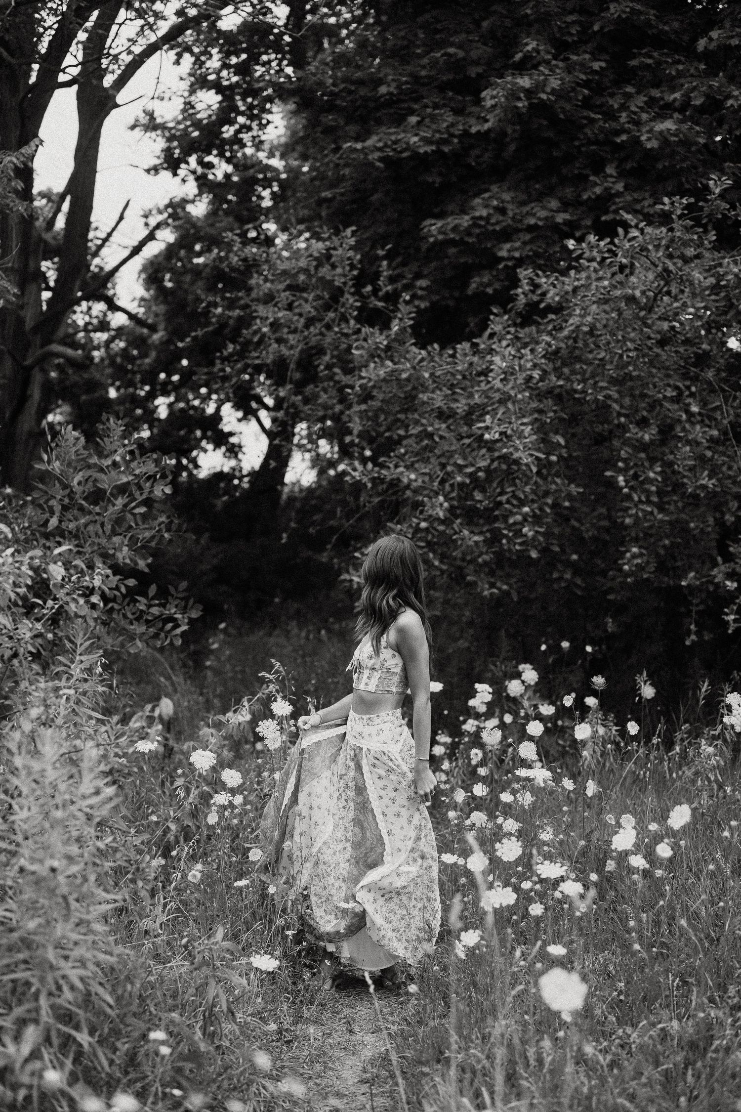 Woman in floral skirt walking through wildflower meadow near trees, black and white photography.