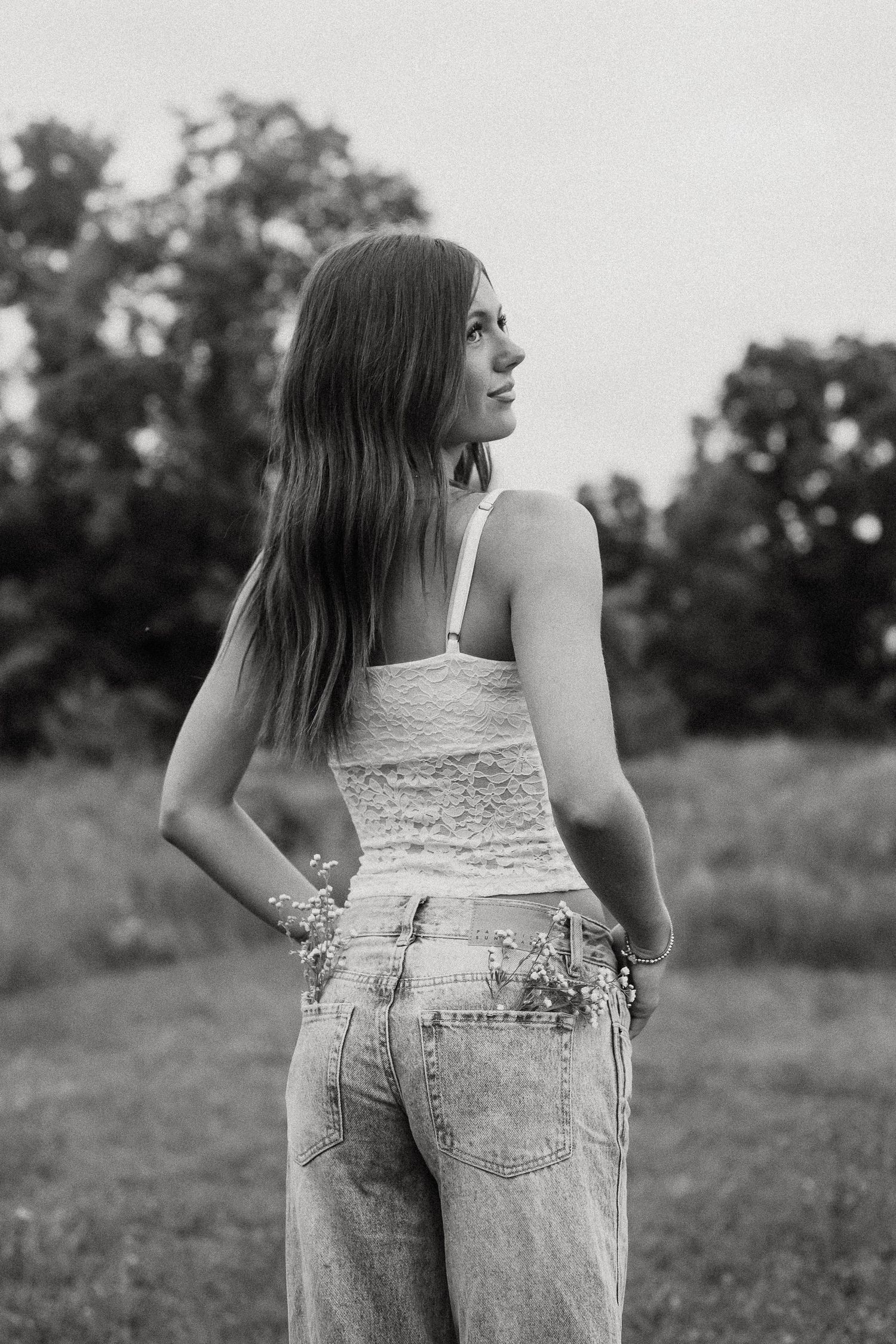Black and white photo of a young woman in lace top and jeans standing in a field with wildflowers.