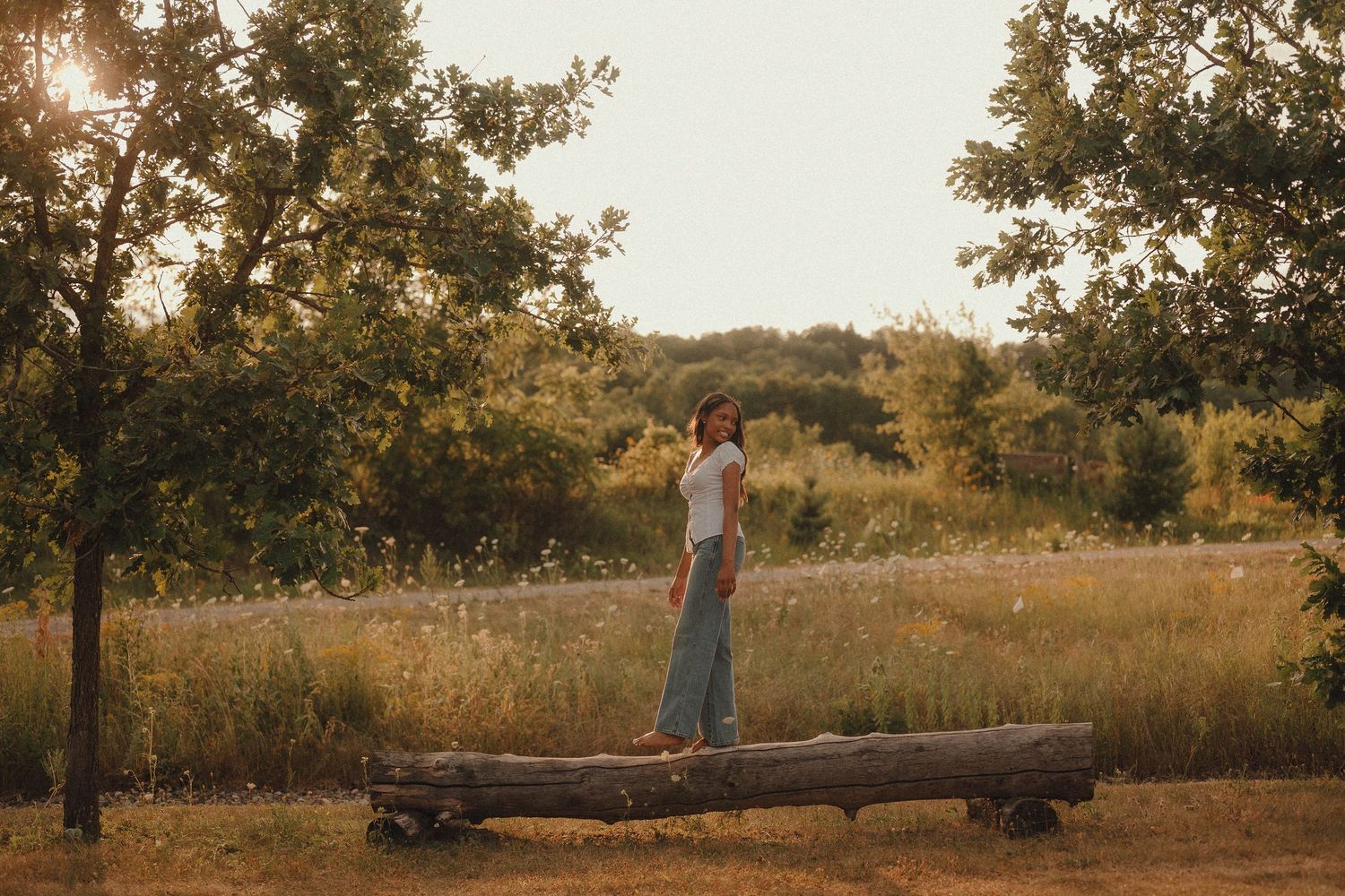 Woman standing on a fallen log in a sunlit meadow surrounded by lush green trees during golden hour.