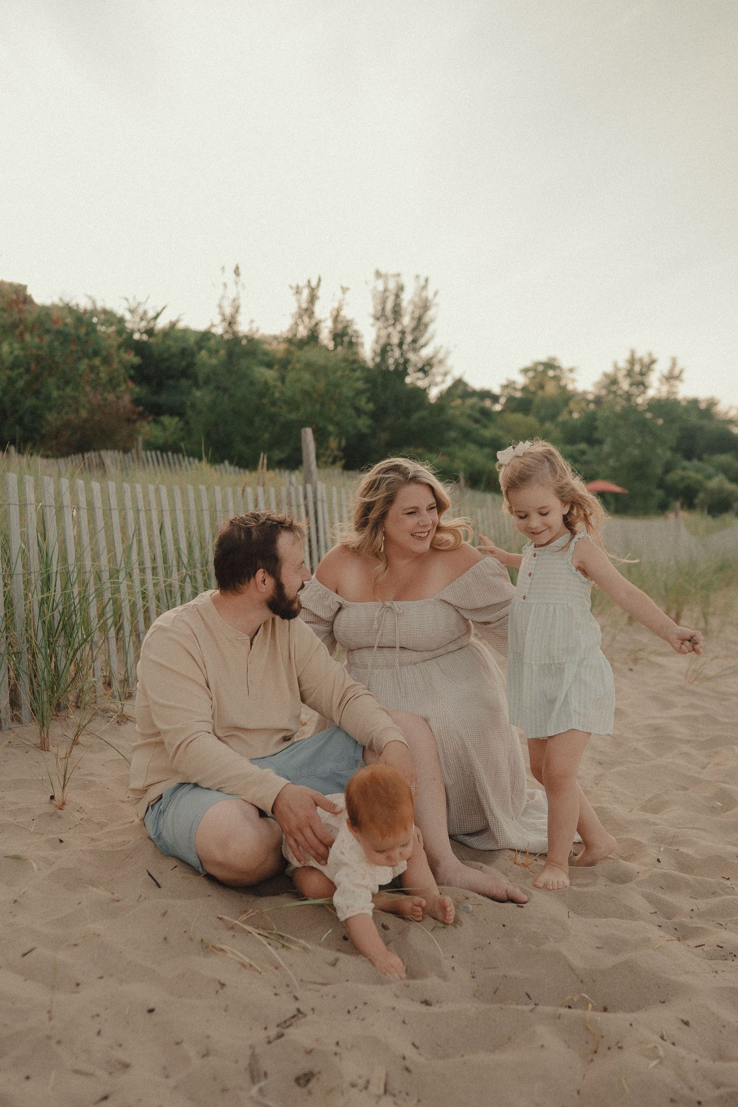 Family of four sitting together on sandy beach dunes near a wooden fence at sunset.