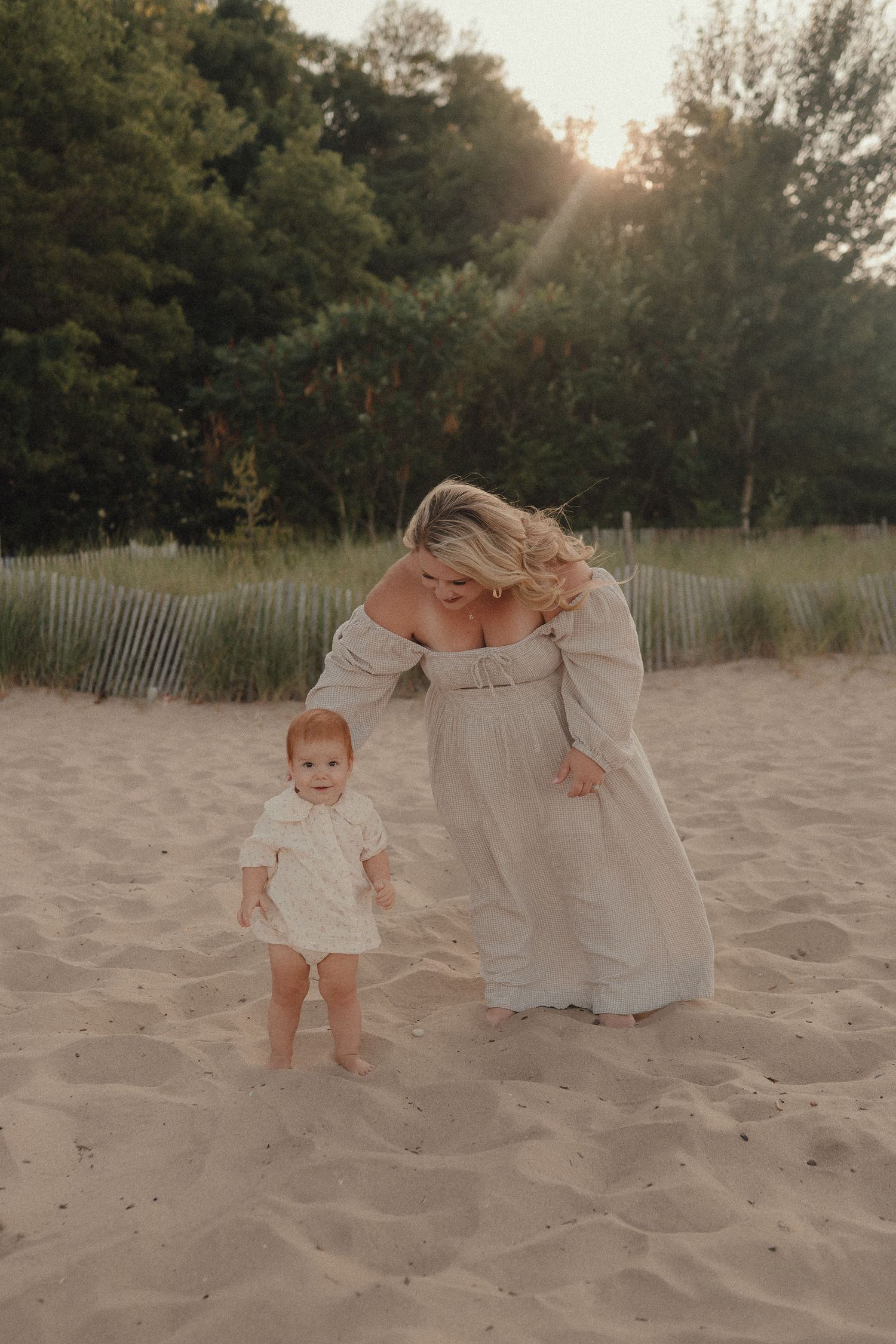 A mother in a flowing dress helps her baby walk on sandy beach dunes at golden hour.