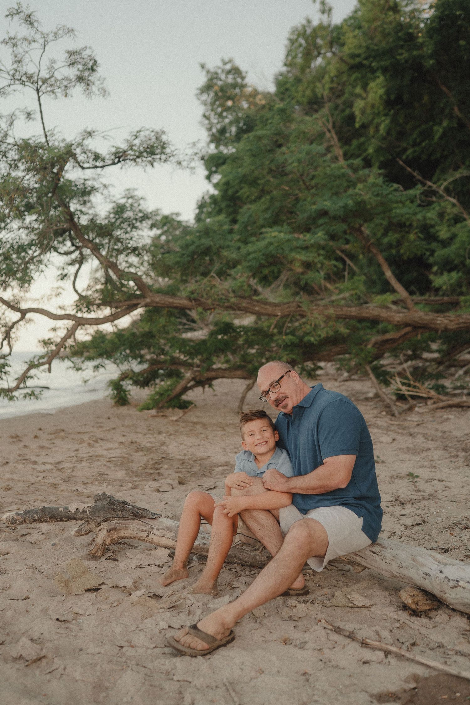 A father and young son share a warm embrace sitting on driftwood at a sandy beach with lush green trees.