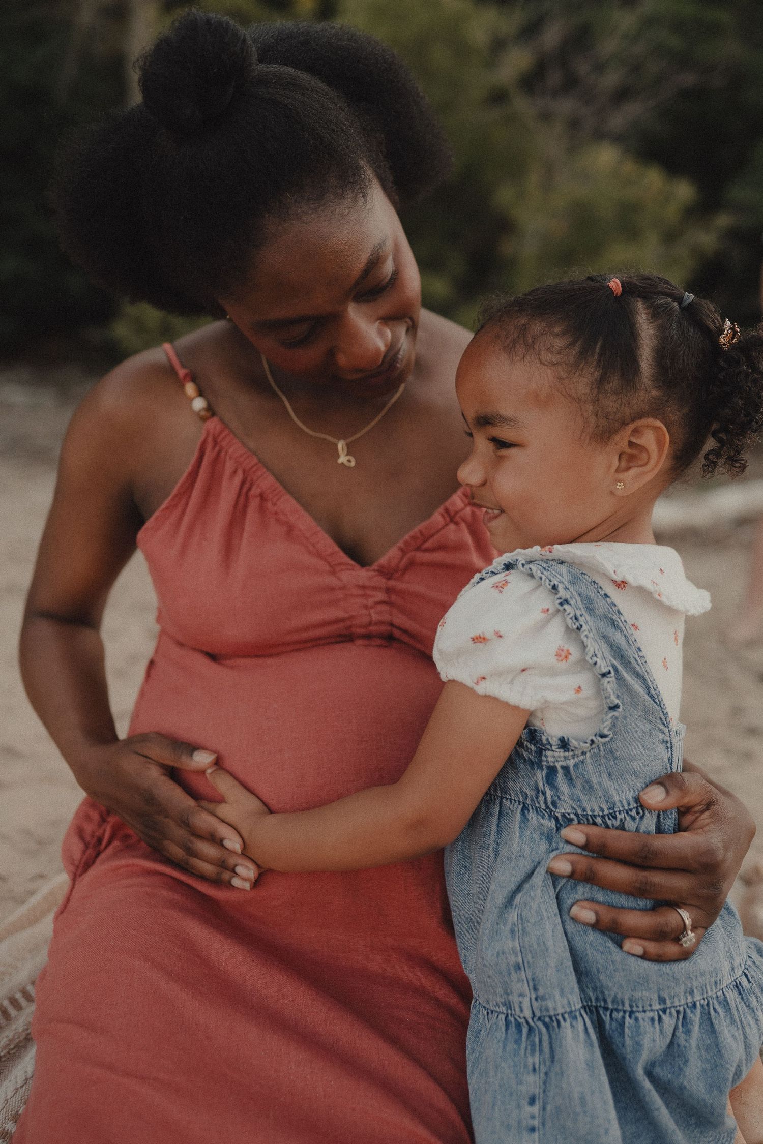 A pregnant Black woman in coral dress tenderly embraces her smiling toddler daughter on a beach.