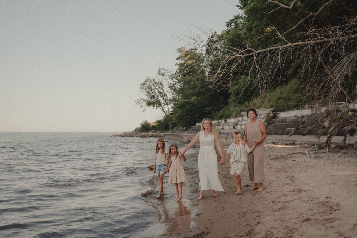 Family of five walks along a sandy shoreline with trees and calm water nearby.