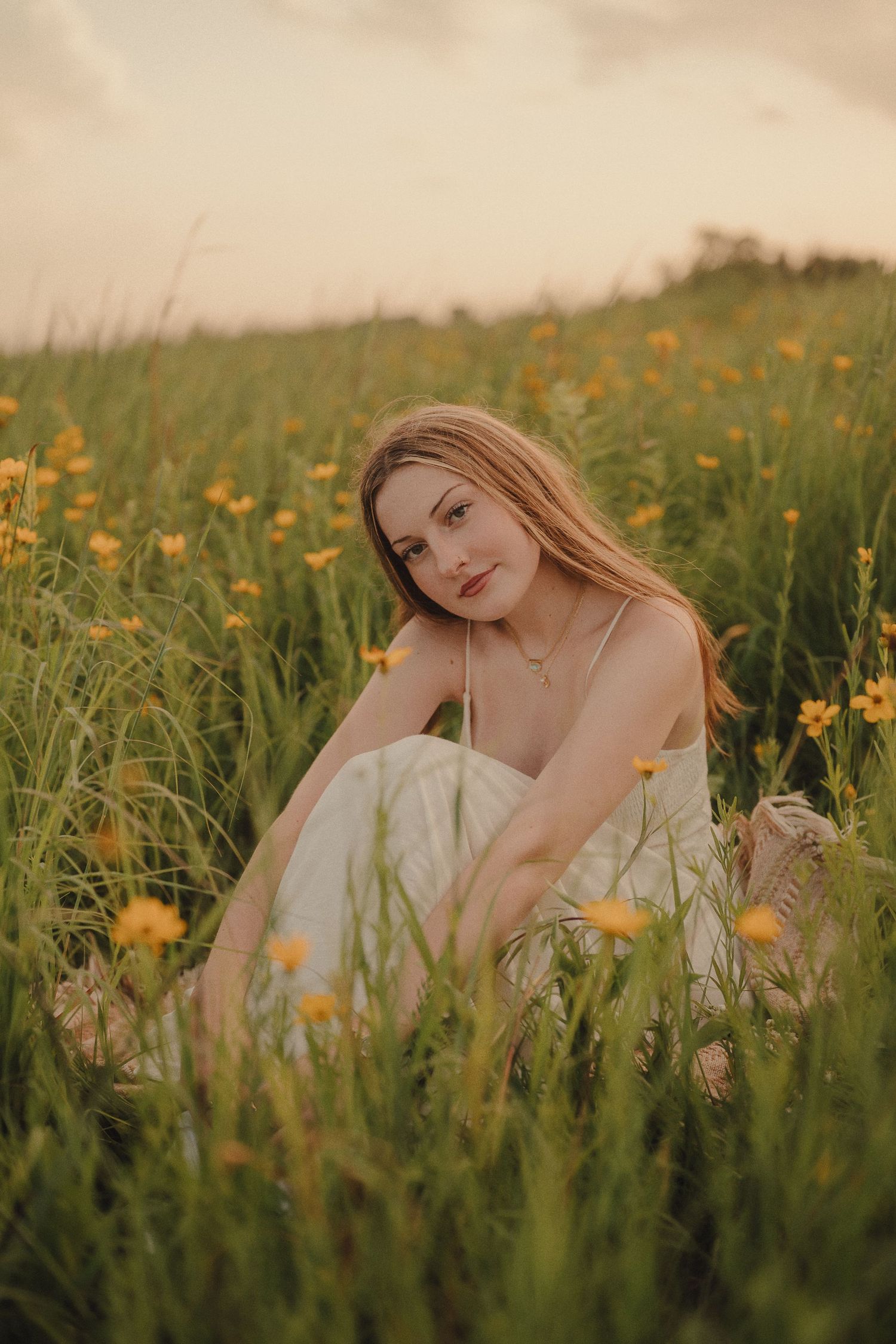 Young woman in white dress sitting among tall green grass and yellow wildflowers at golden hour.
