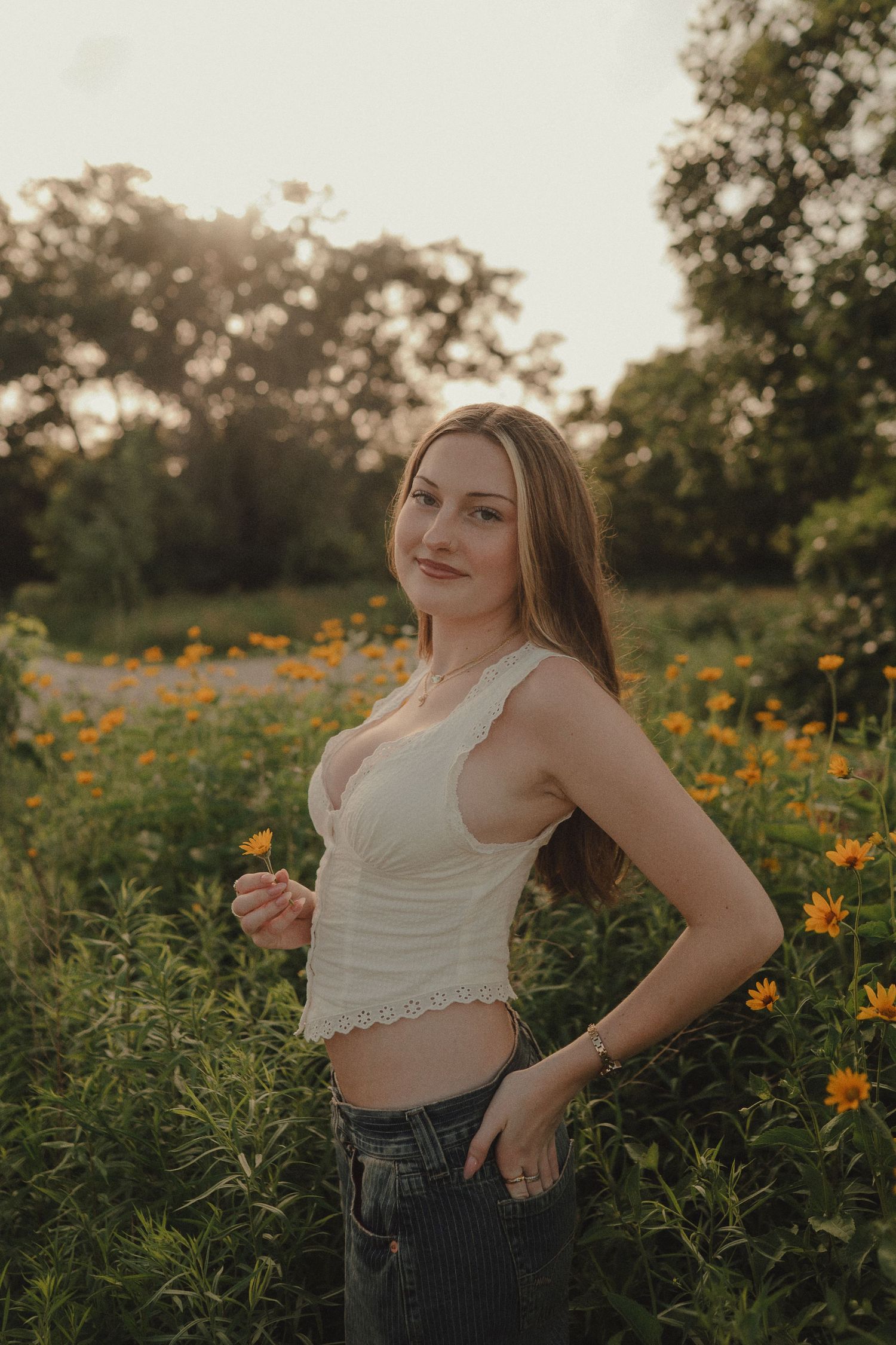 Young woman in white crop top holding a yellow flower while standing in a vibrant field of orange blooms at sunset.
