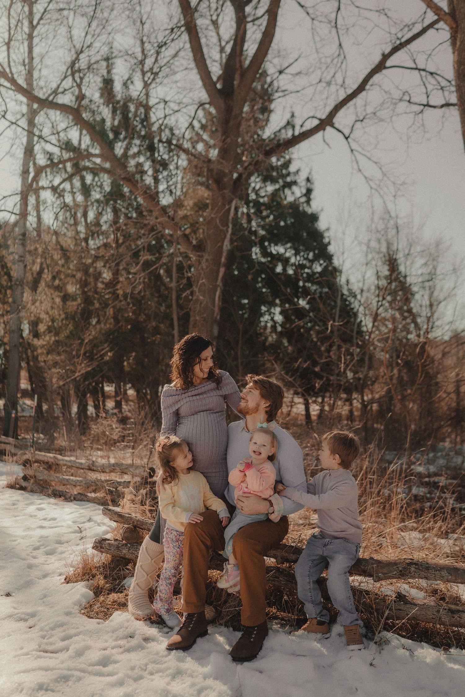A family sits together on a snowy log in a winter forest setting with bare trees in the background.