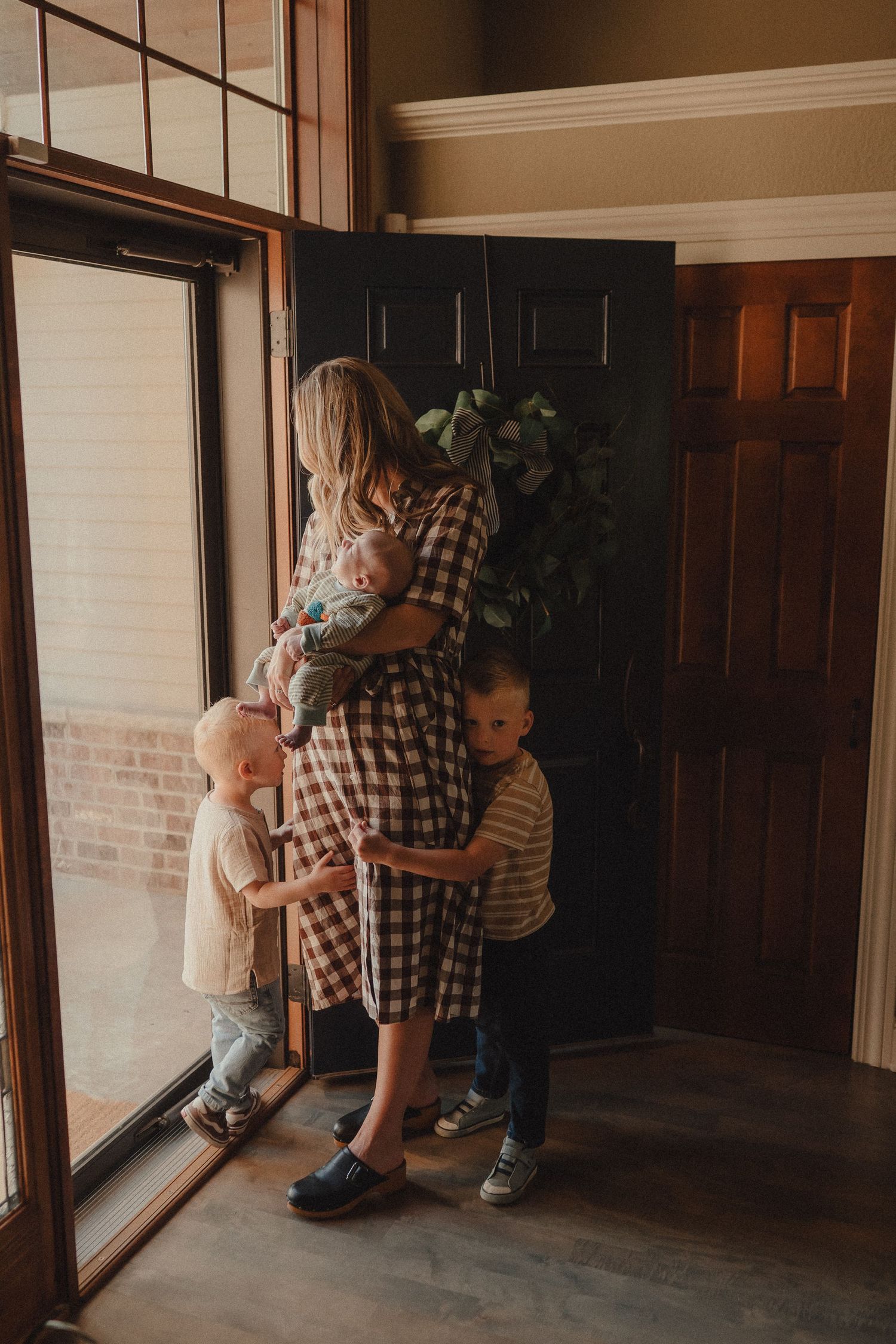 A mother in a plaid dress holds her newborn while two young boys hug her near the front door.