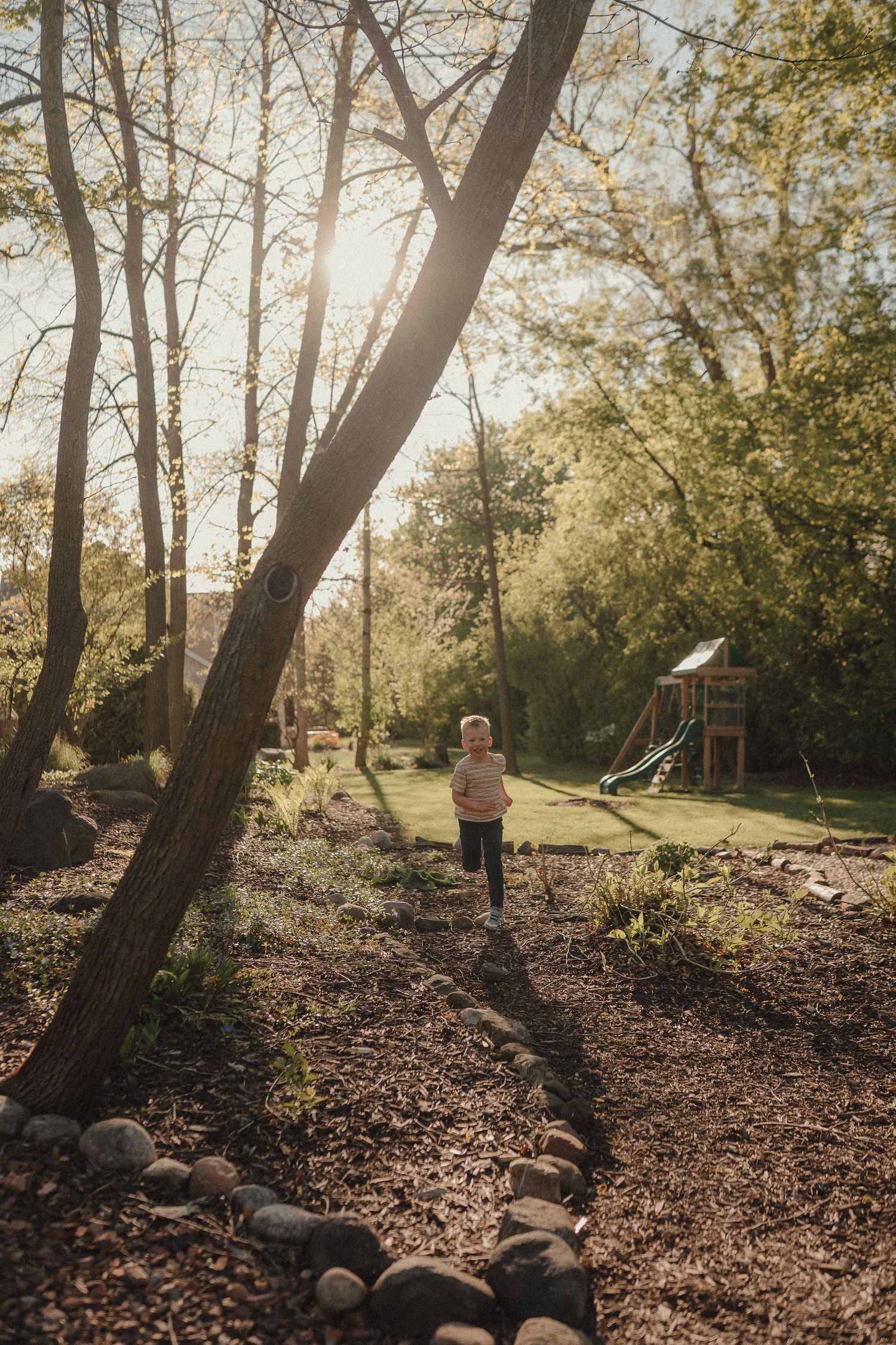 A young child runs along a mulch path bordered by rocks in a sunlit backyard with trees and a playground.