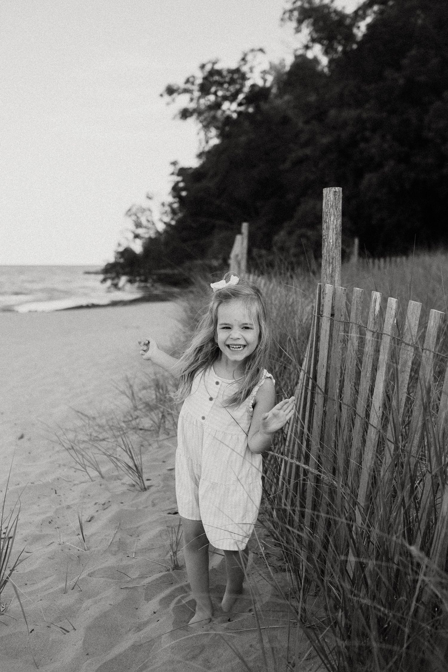 Happy young girl in white romper smiling beside a wooden beach fence in black and white photo.