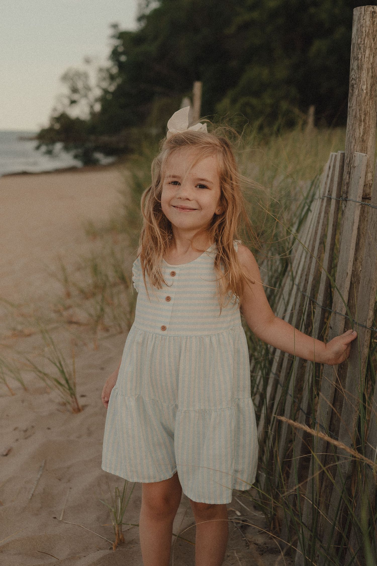 Young girl in striped romper smiling on sandy beach path beside wooden fence.