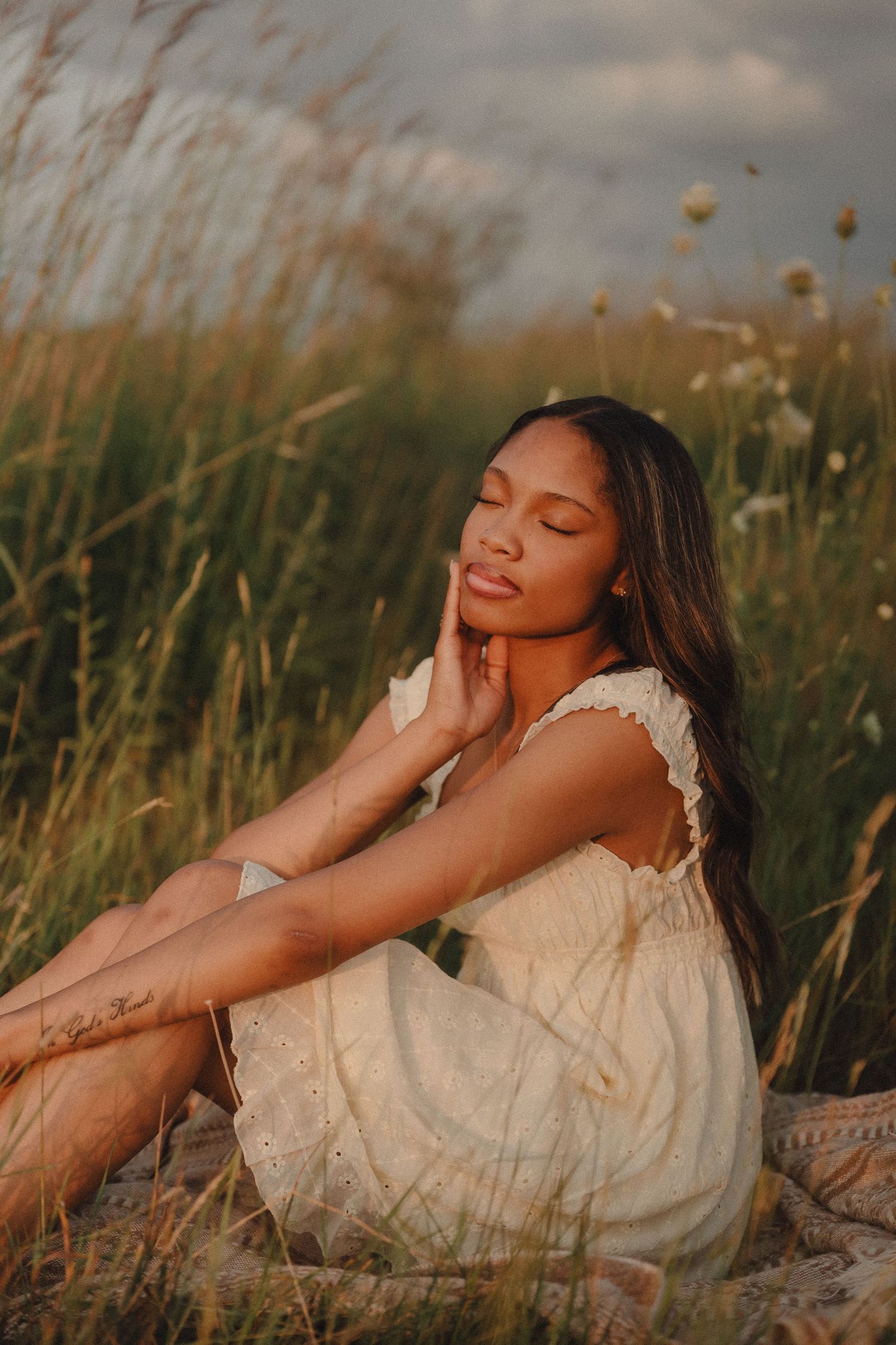 Young woman in white lace dress sits peacefully in tall grass field at sunset with eyes closed.
