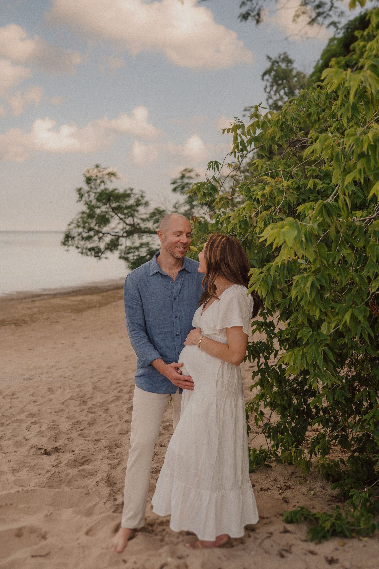 A romantic couple embraces on a beach with lush green foliage and cloudy skies in the background.