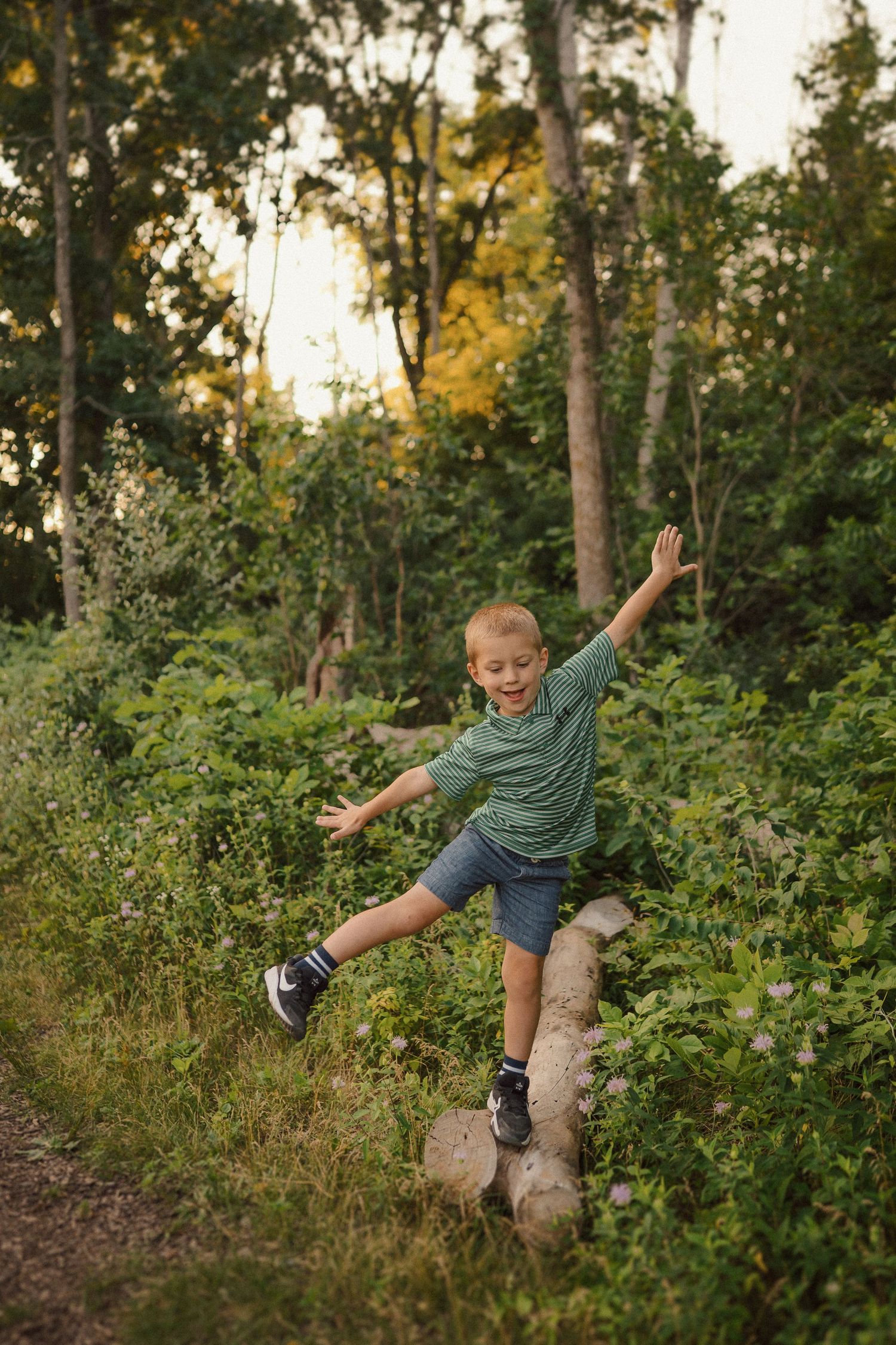A young child strikes a playful pose while balancing on a rock in a wooded forest setting at sunset.