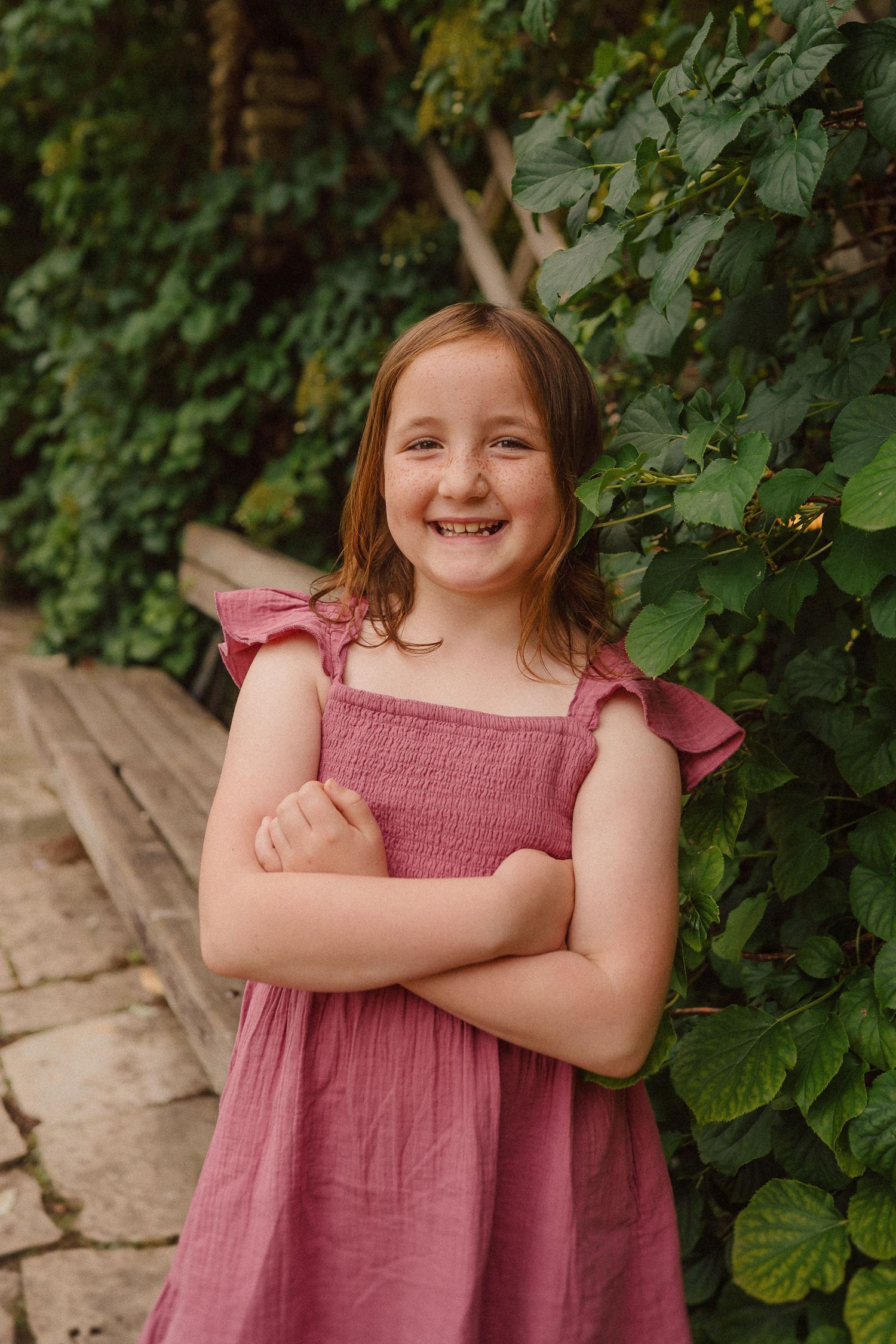 Young child in dusty rose dress poses cheerfully against lush green foliage.