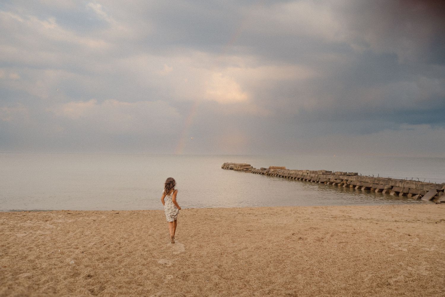 Person in white dress walking alone on sandy beach near wooden pier under cloudy sky.