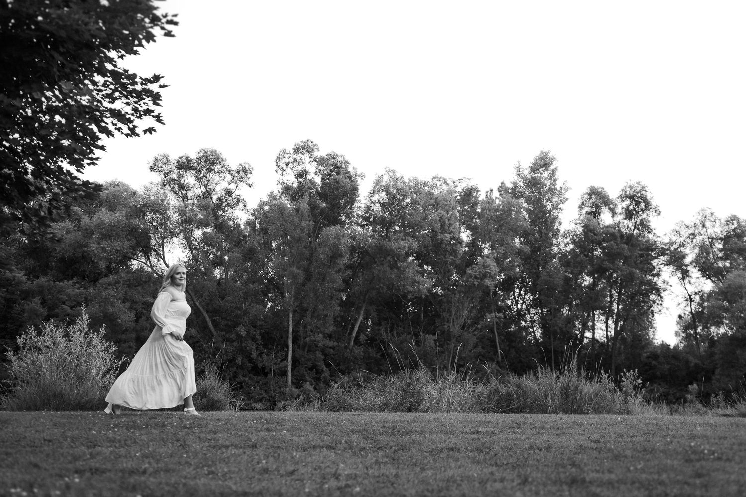 Person in flowing dress stands atop a grassy hill against a tree-lined horizon at dusk.