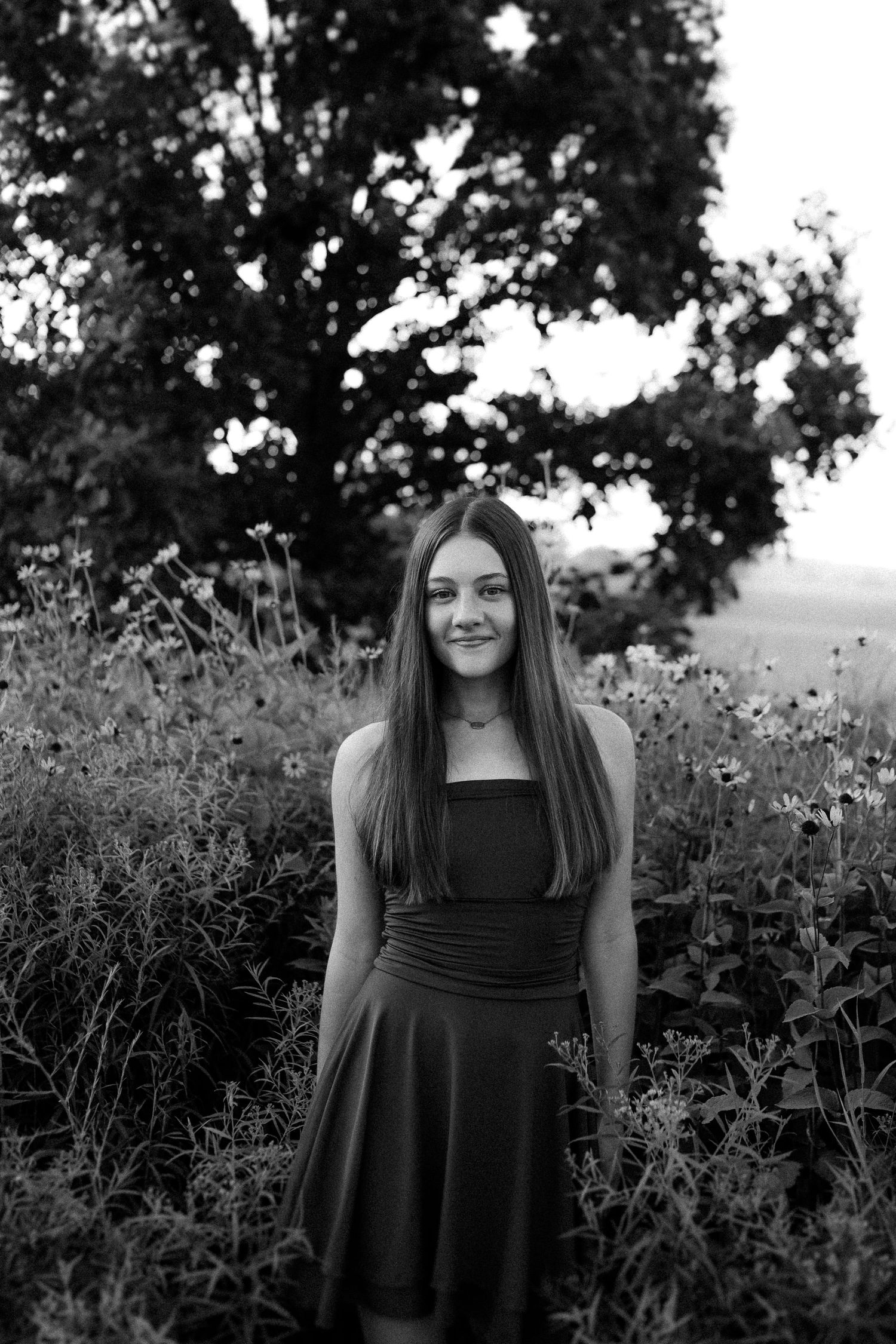 Black and white portrait of someone wearing a sleeveless dress standing in a wildflower meadow at dusk.