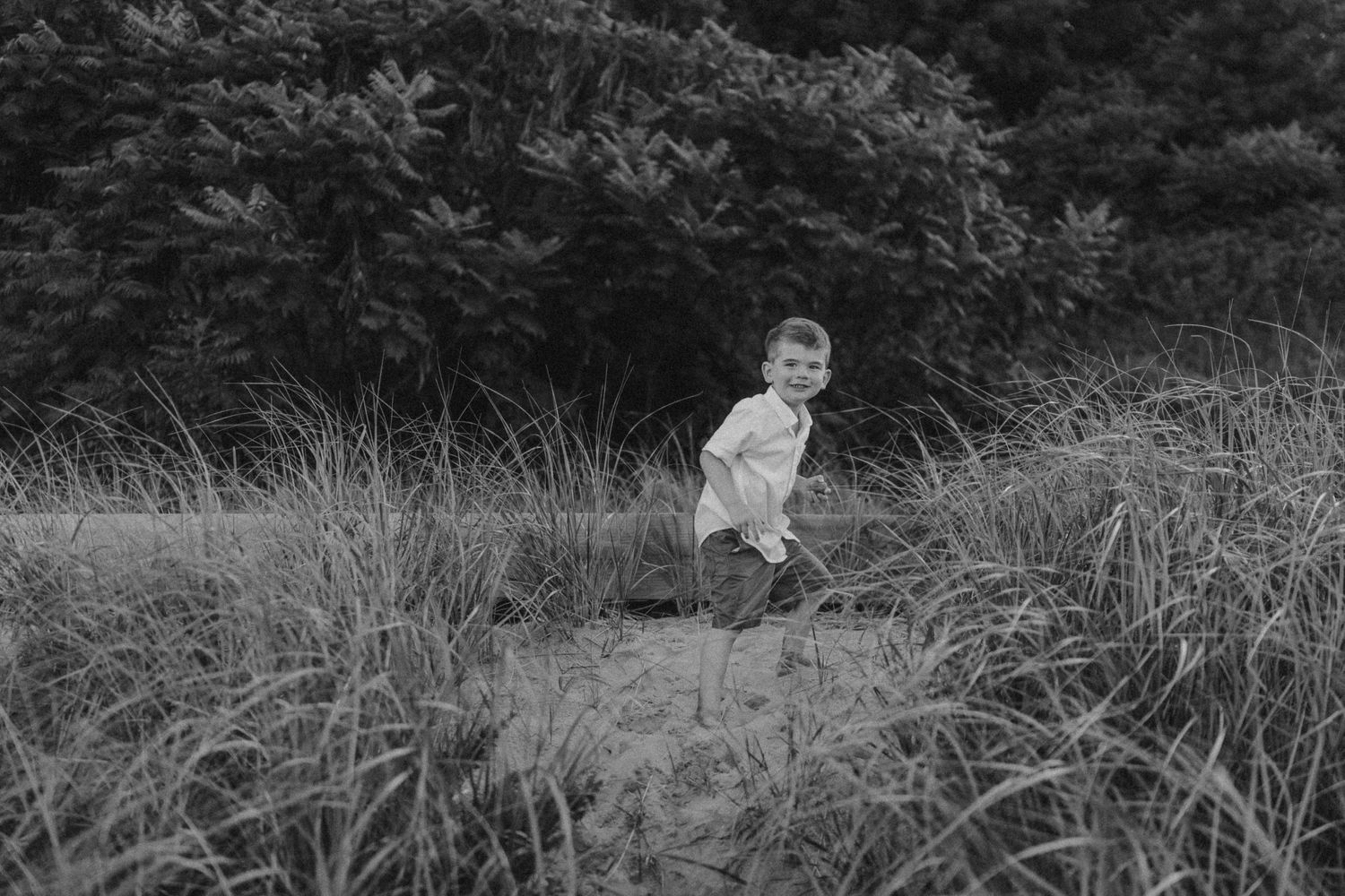 Black and white photo of a toddler walking through tall grass in nature.