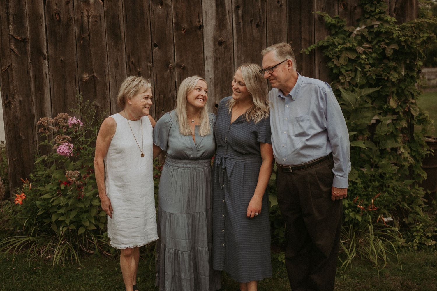 Multi-generational group portrait in casual attire standing against a rustic wooden fence in a garden setting.