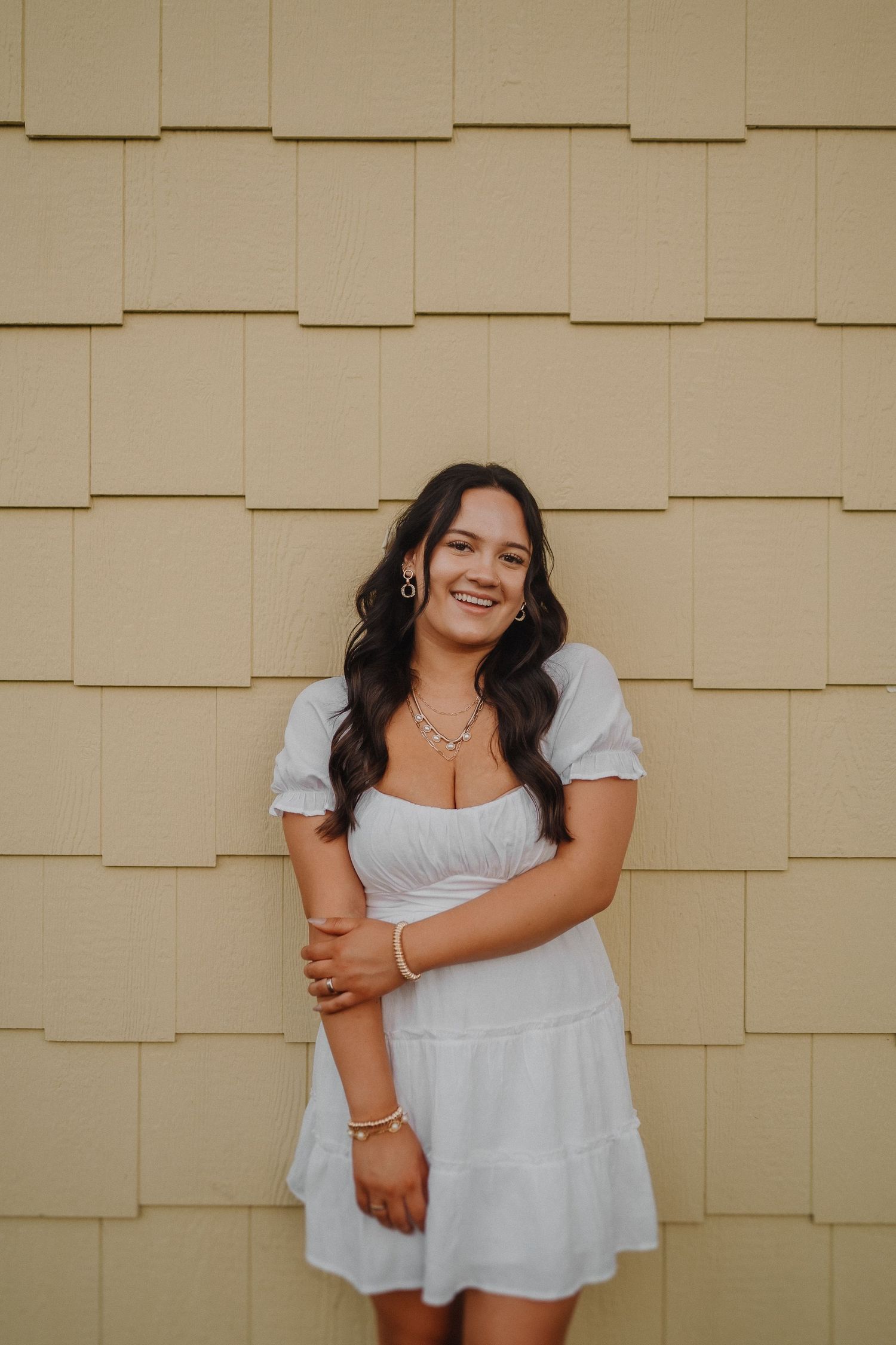 Someone in a white summer dress poses against a beige shingle wall while wearing jewelry and smiling warmly.