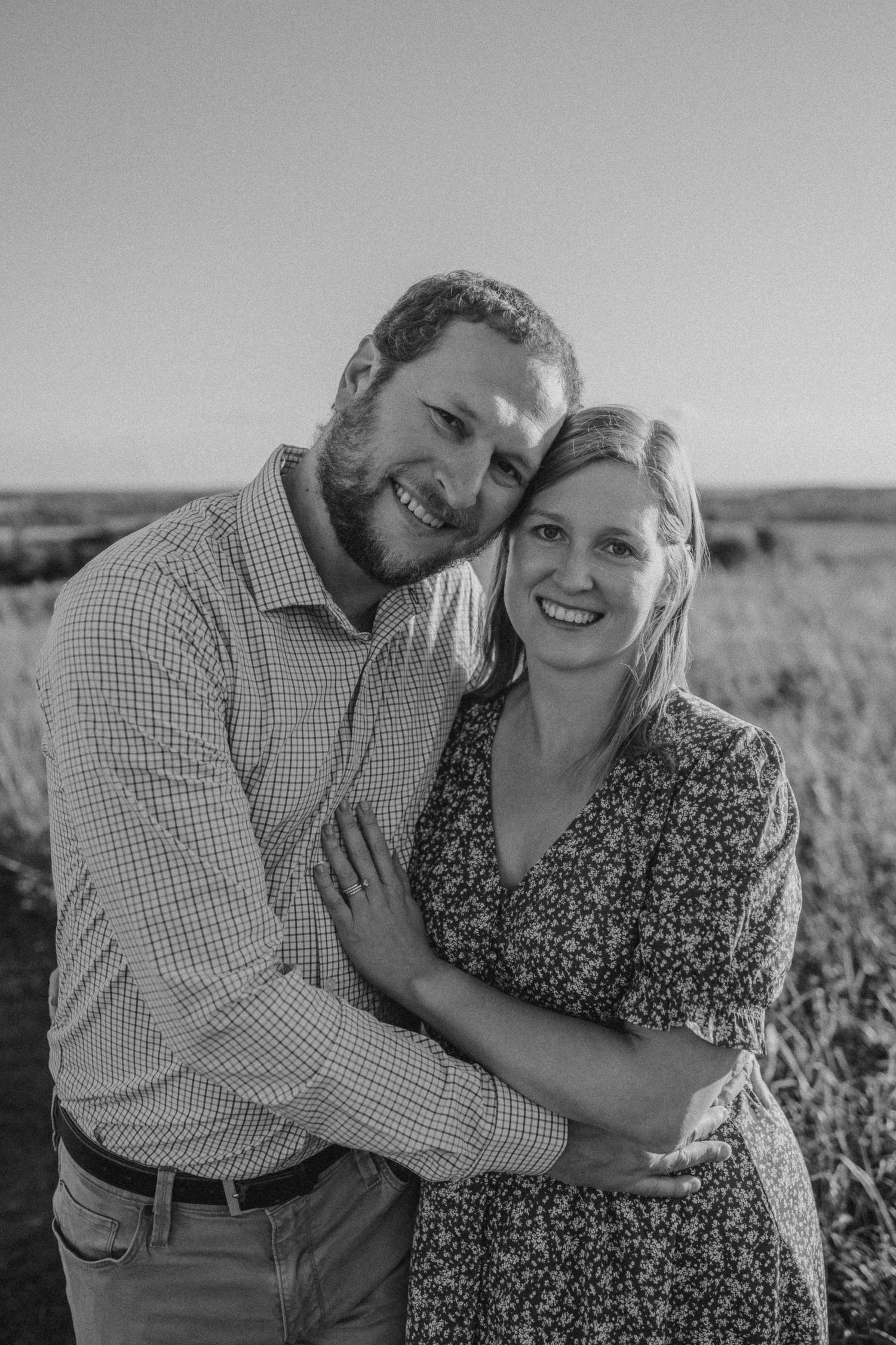 A couple embraces while smiling together in a field during a black and white portrait session.