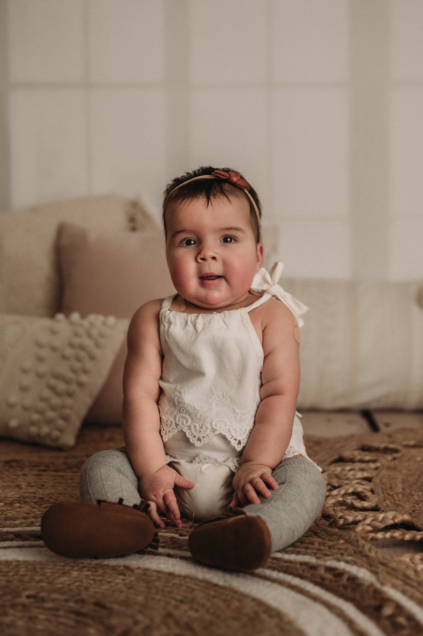 Baby wearing white lace outfit and headband sits on brown patterned rug with decorative pillows.