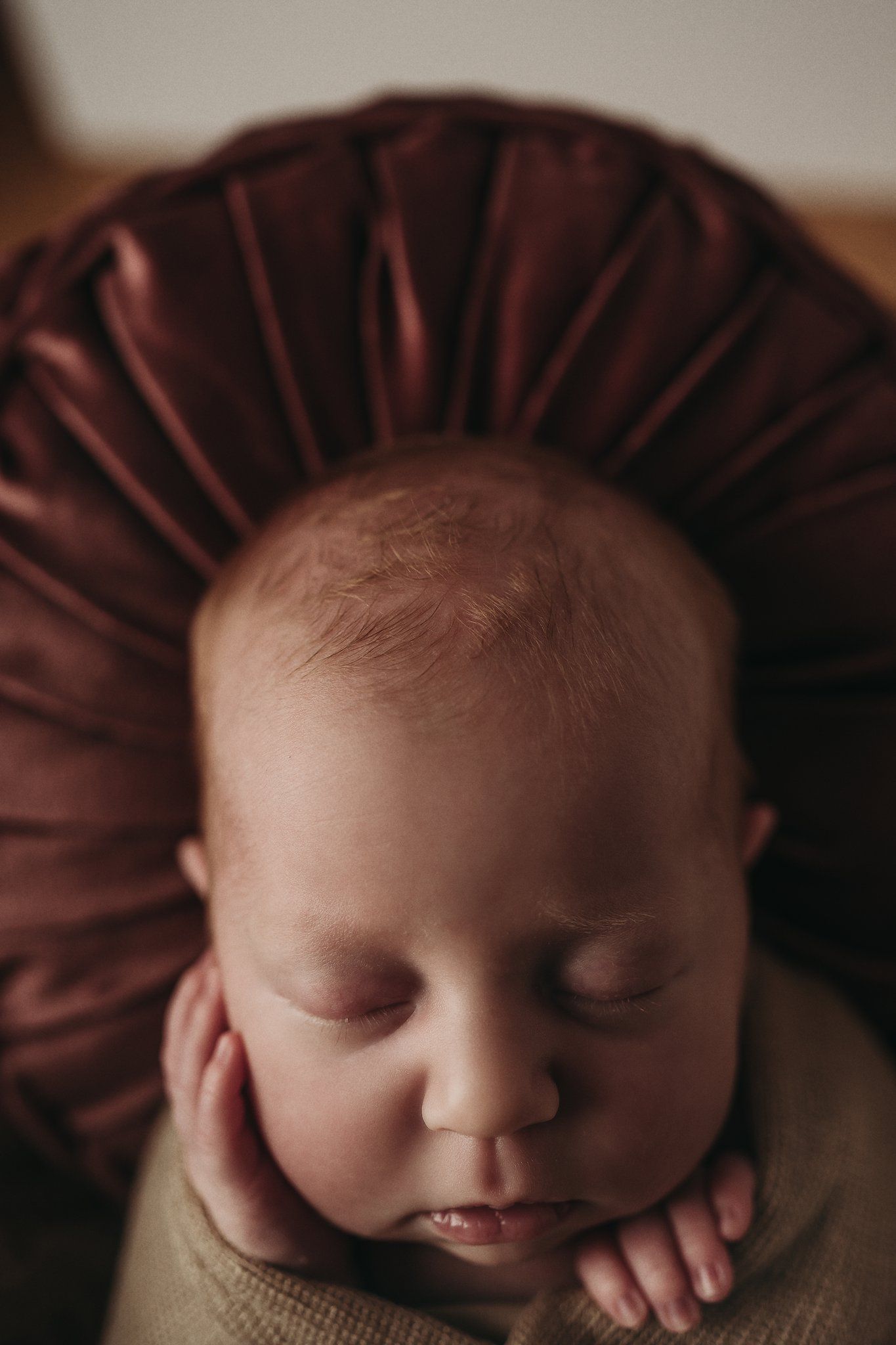 Peaceful sleeping infant posed against burgundy circular cushion wearing neutral wrap.
