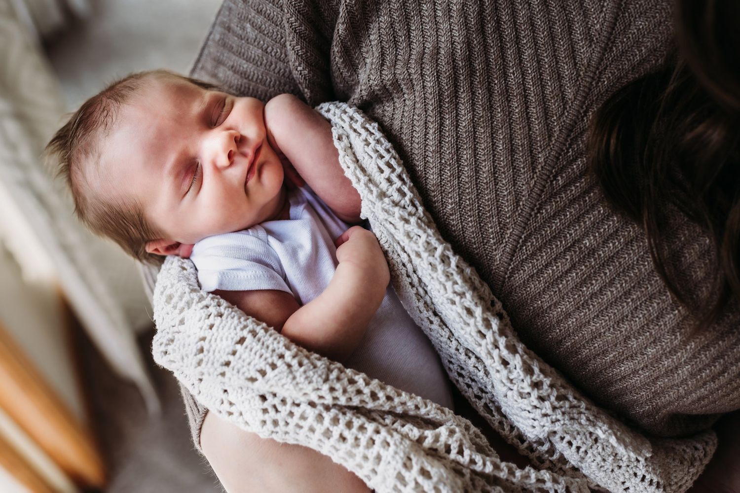Peaceful sleeping newborn swaddled in cream knit blanket against brown sweater.