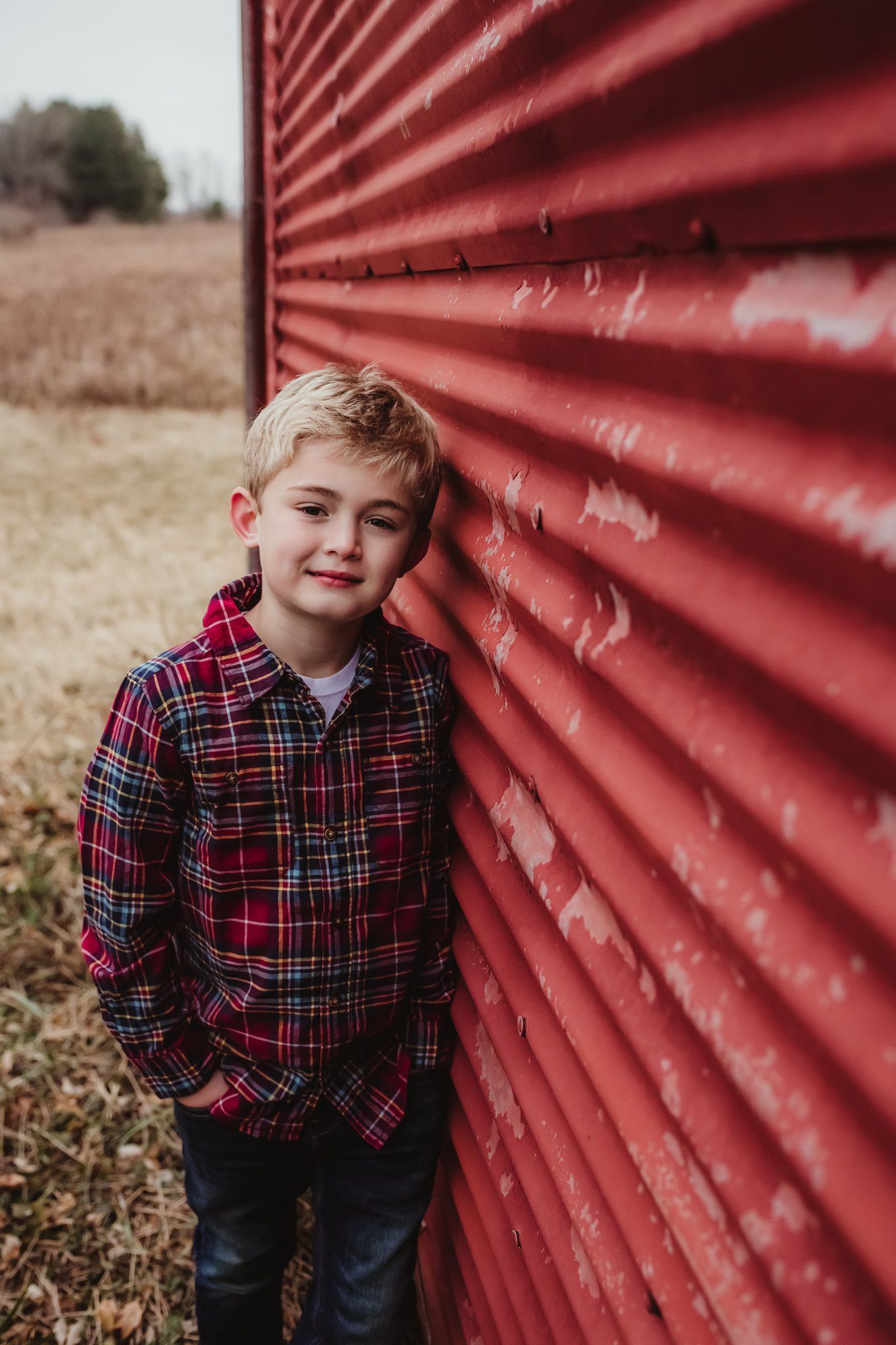 A child in a plaid flannel shirt poses against a red corrugated metal wall in a rustic outdoor setting.