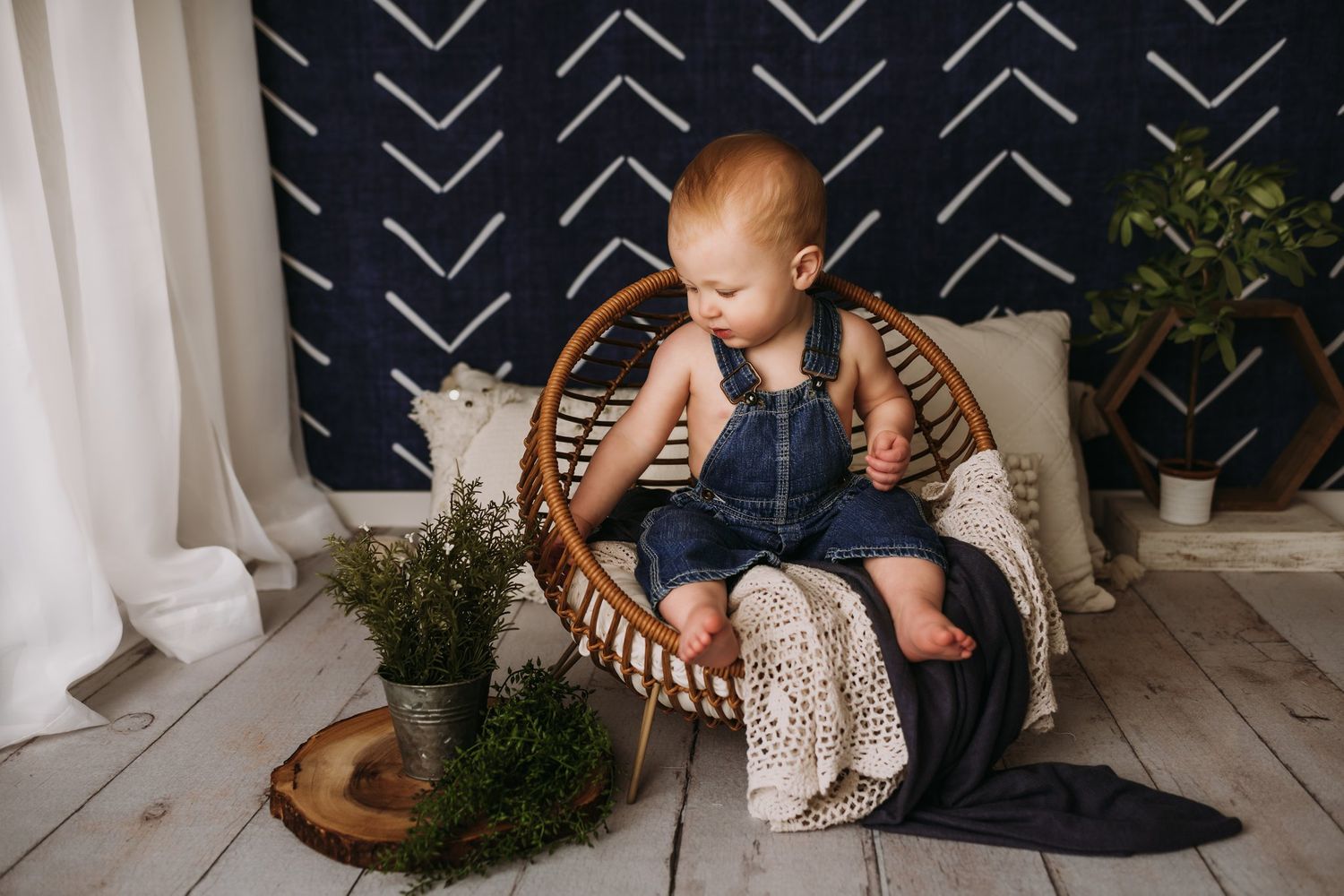 Baby in denim overalls sits in round wicker chair against geometric patterned wall.