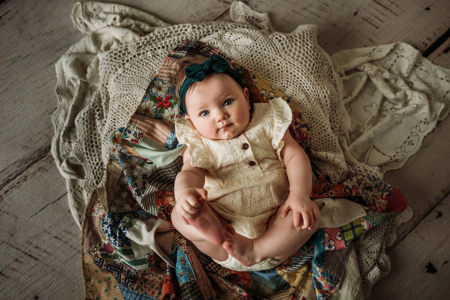 Baby wearing cream colored romper rests on vintage lace and floral fabric in professional photo shoot.
