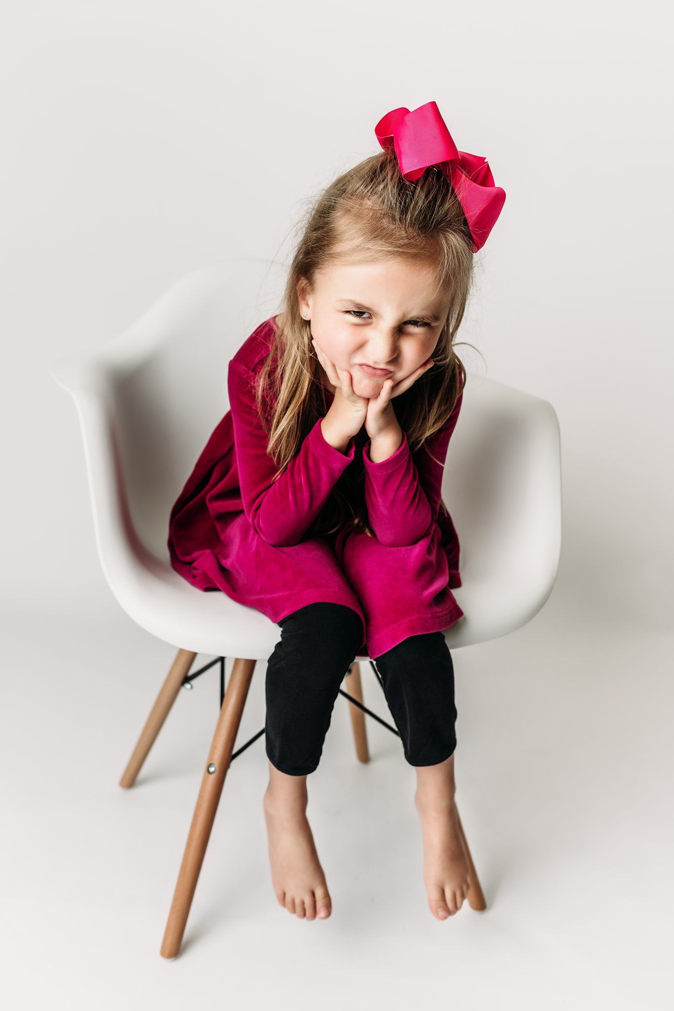Child in burgundy dress poses playfully on white chair with hand on cheek.