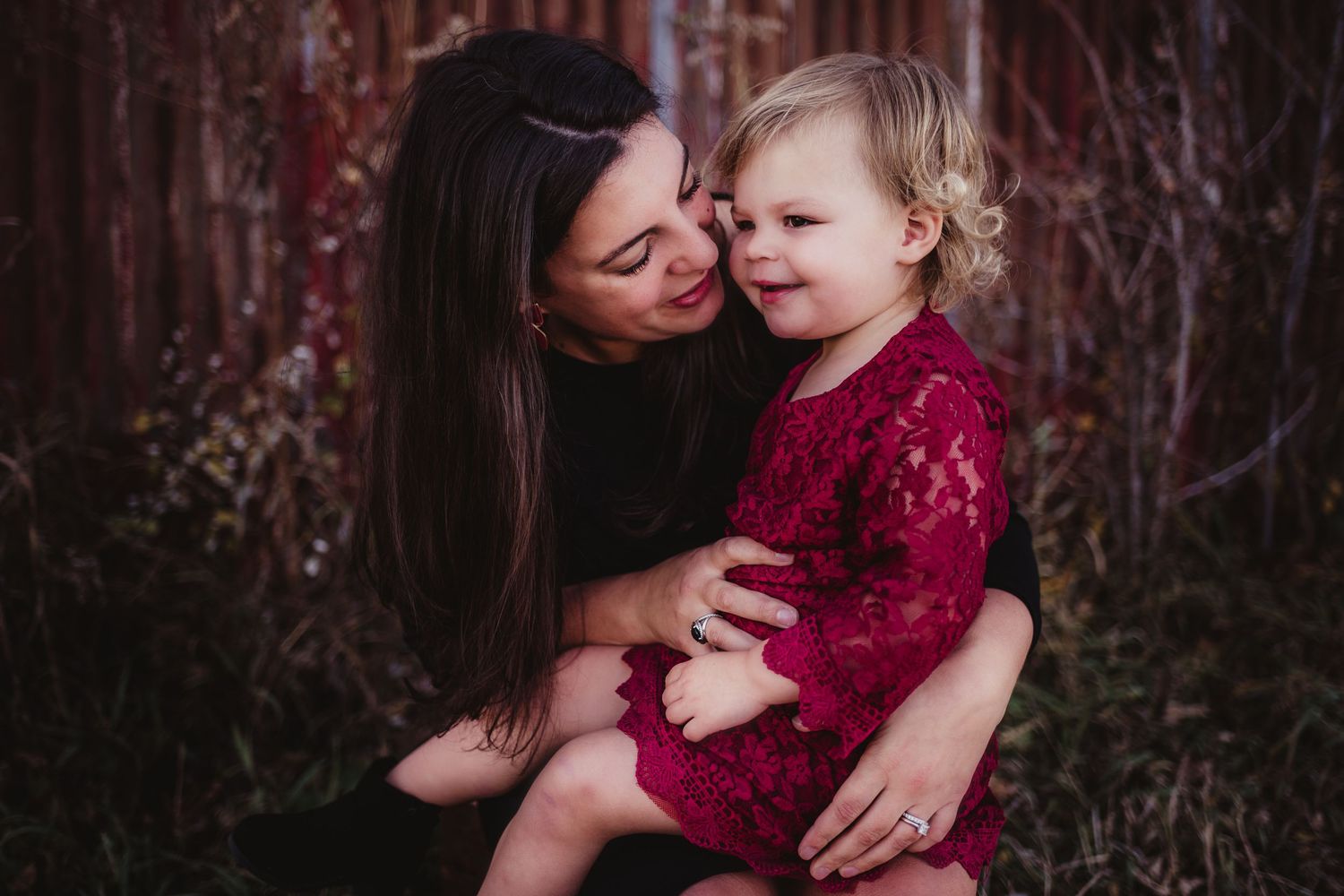 Tender moment between parent and child wearing red dress against rustic background.