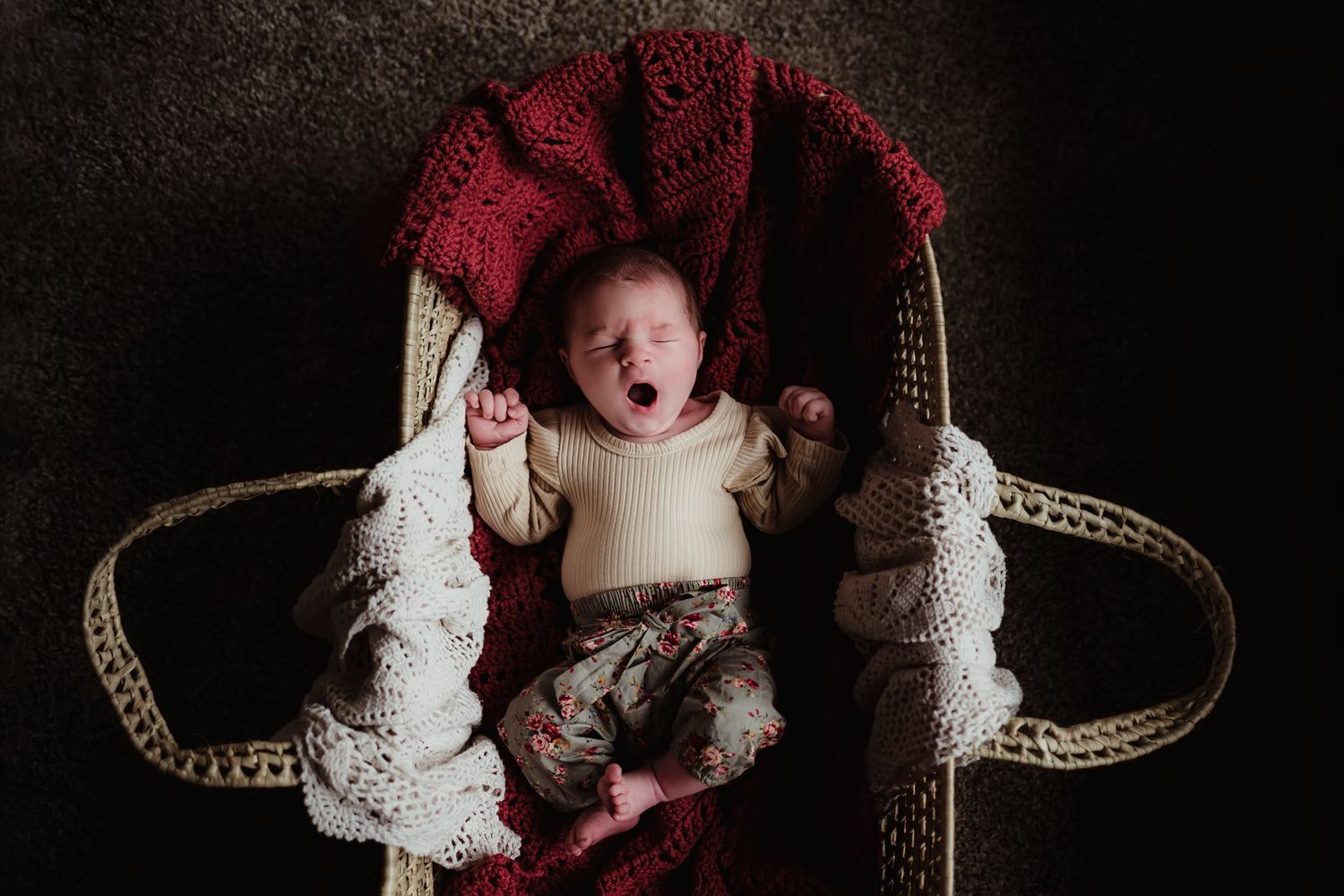 A smiling baby lies on a woven basket with red and white decorative elements against a dark background.