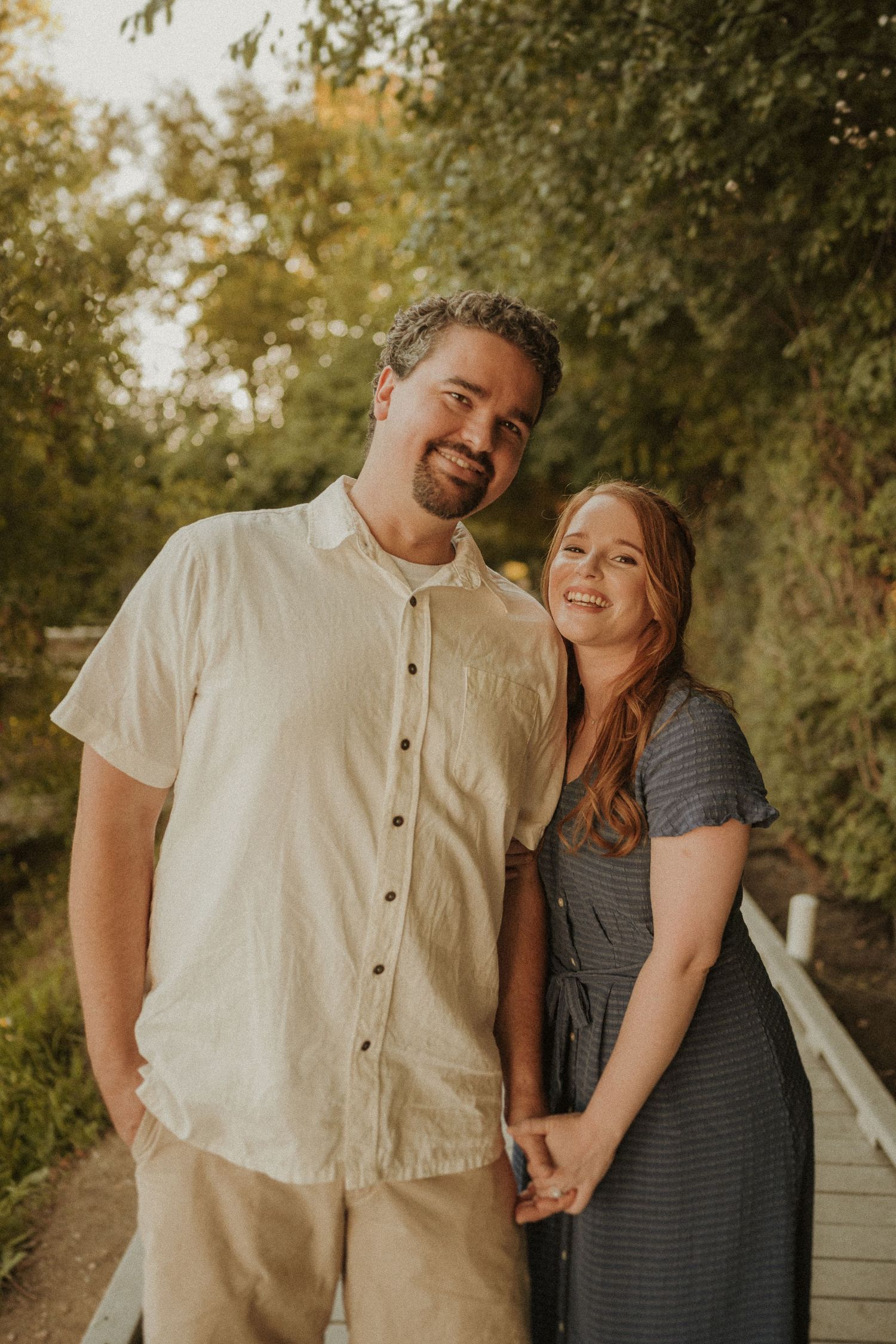 Happy couple stands close together on wooden walkway surrounded by trees in golden afternoon light.
