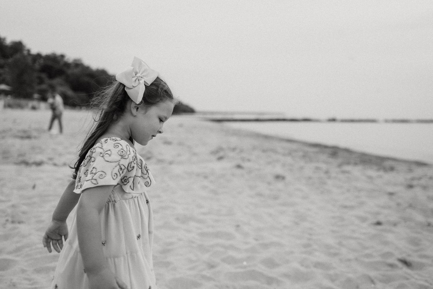Black and white portrait of young child in dress looking down at sandy beach.