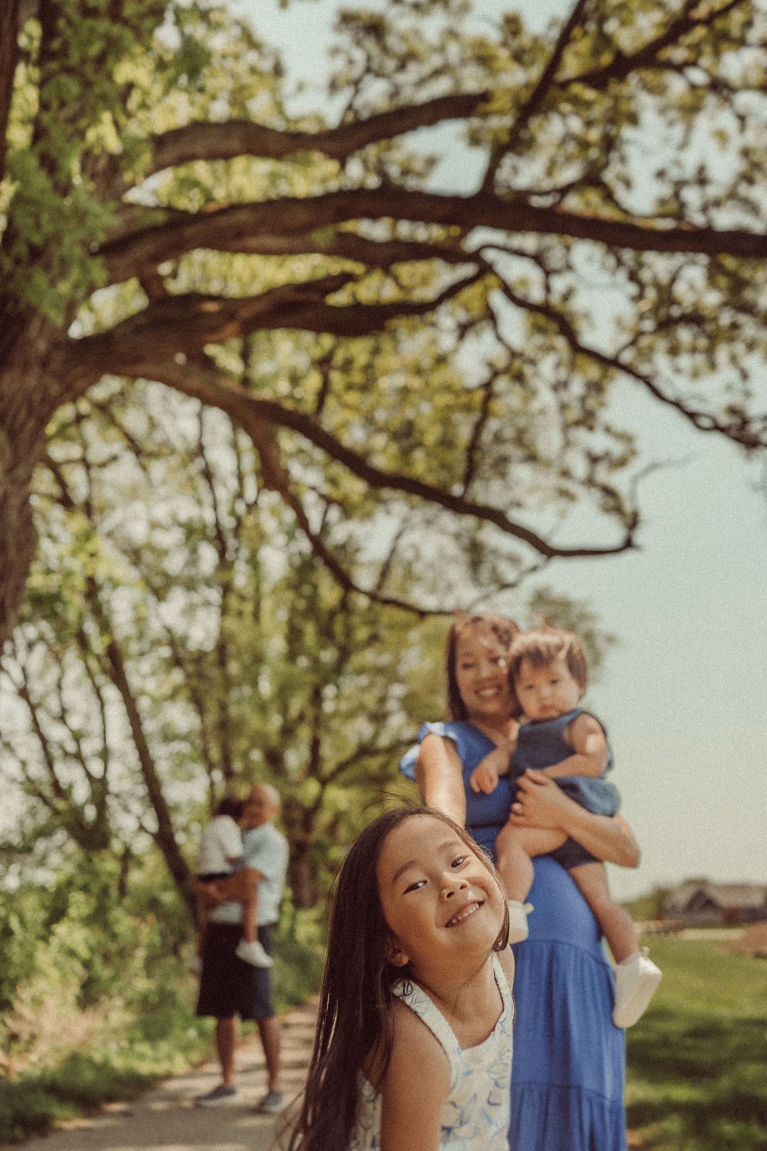 Young girl looking up at tree branches with family members in background on nature path.
